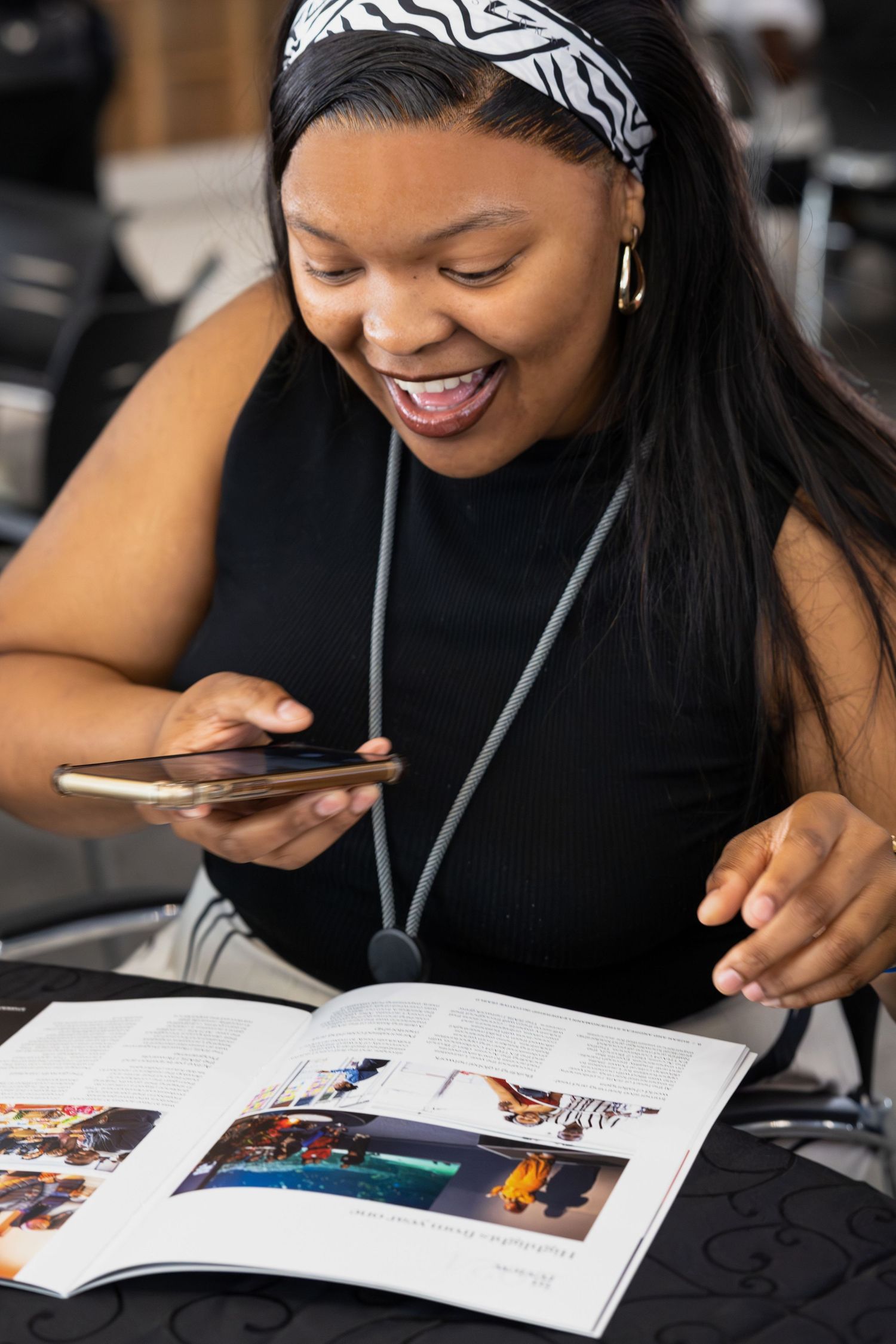 Smiling person in black top looks at phone while reading magazine at desk.