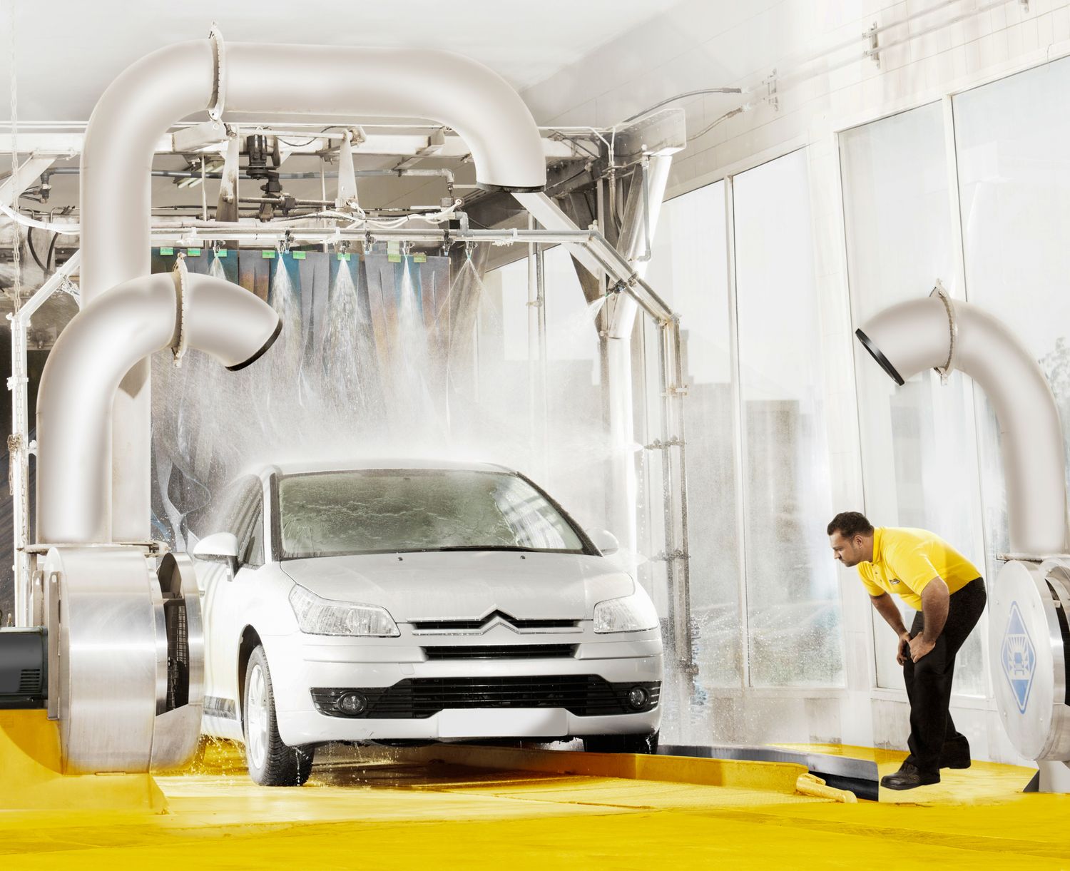 White car going through an automated car wash system with large mechanical washing equipment and water sprayers.
