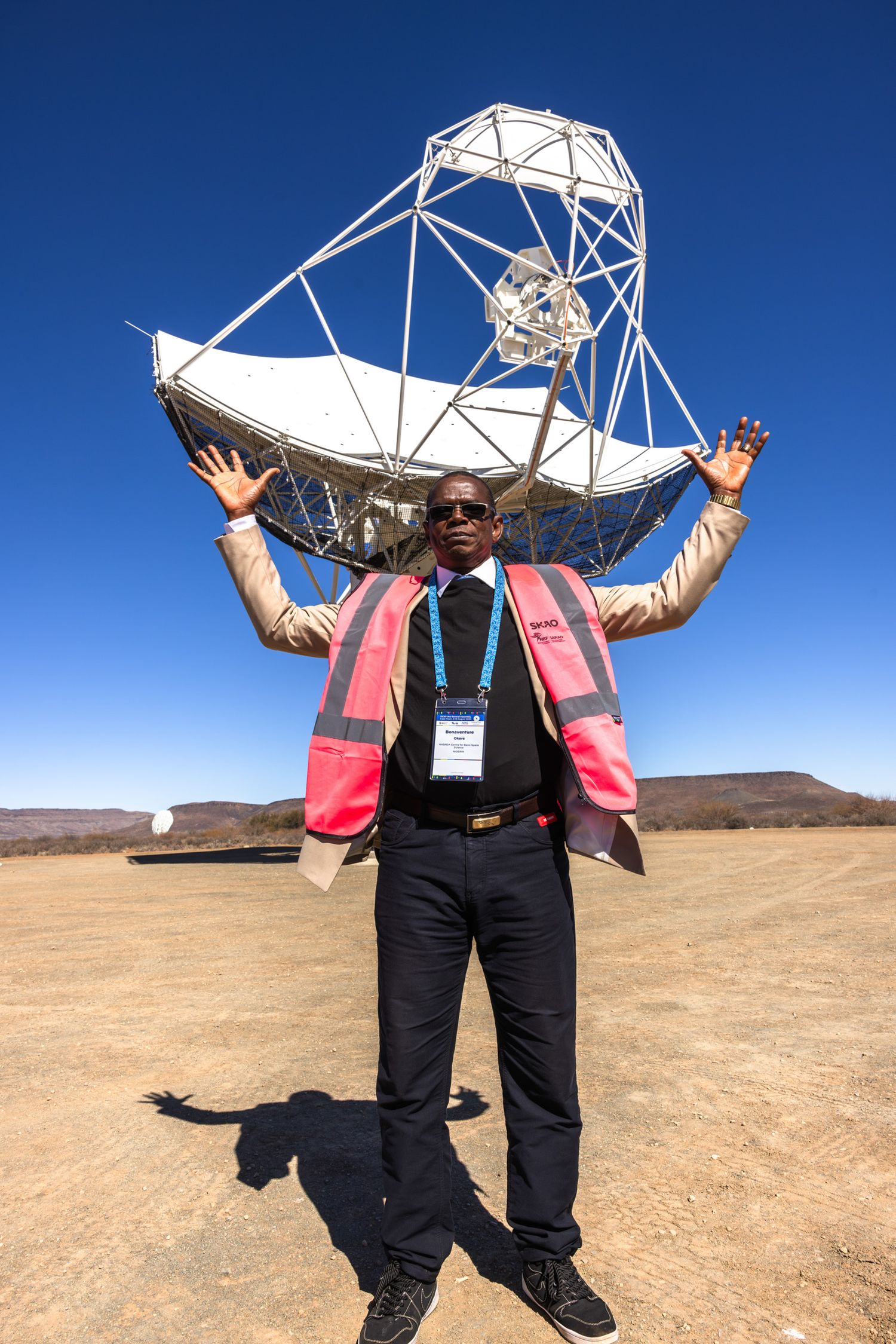Person poses enthusiastically in front of large satellite dish in desert setting.