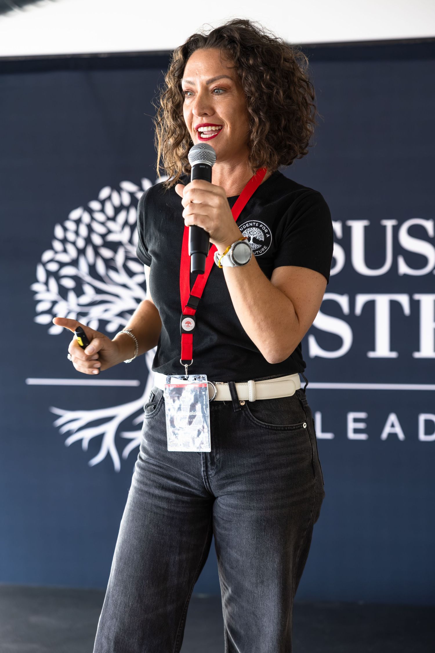 A speaker wearing a black top and red lanyard gestures energetically while presenting at a business event.