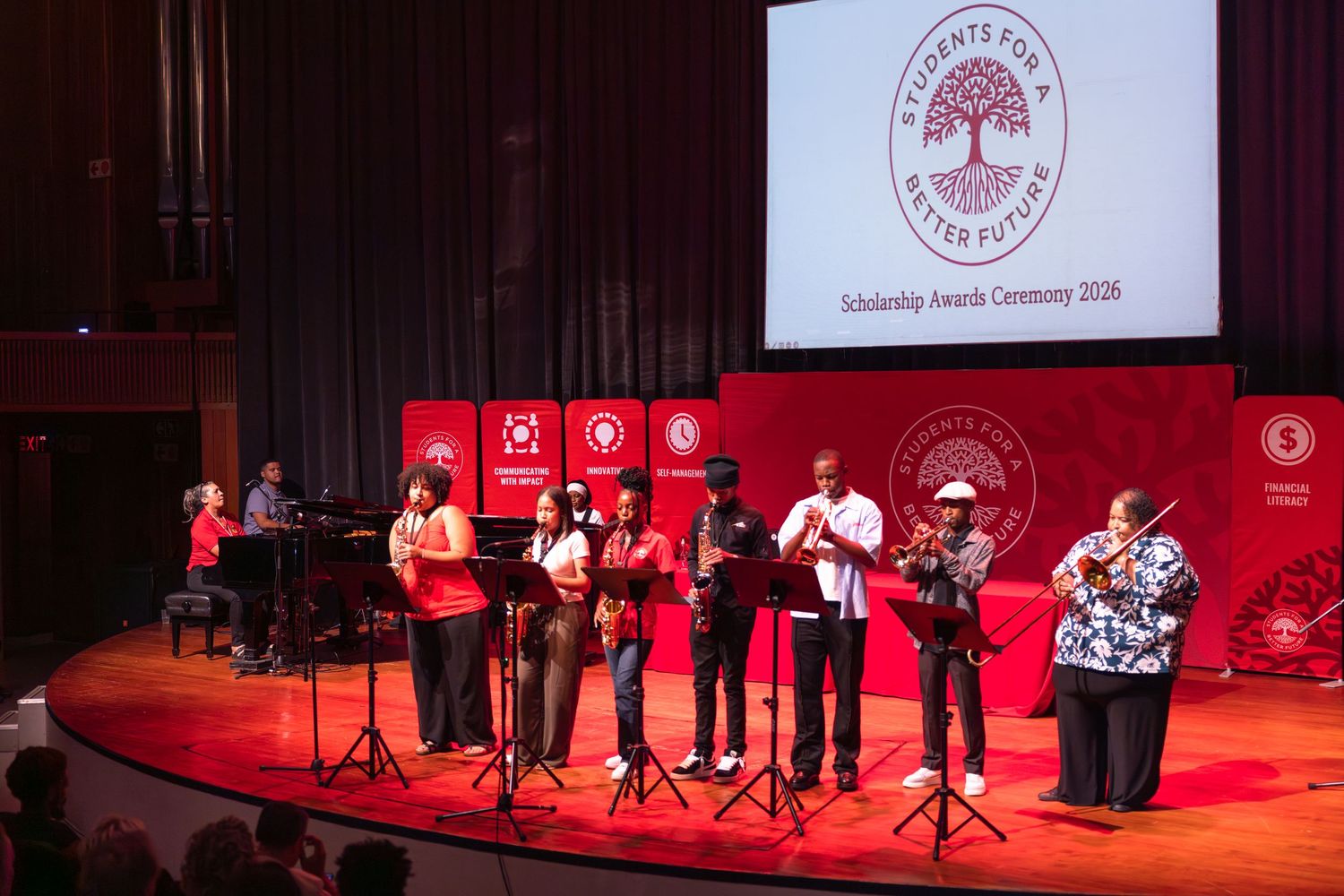 Musicians perform on stage at a concert with red lighting and a university logo projected on screen behind them.