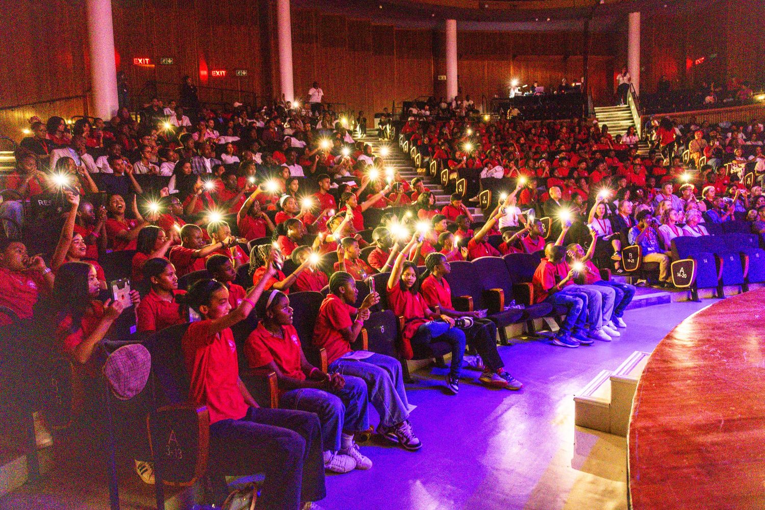Large audience in auditorium illuminated by dramatic purple and red stage lighting.