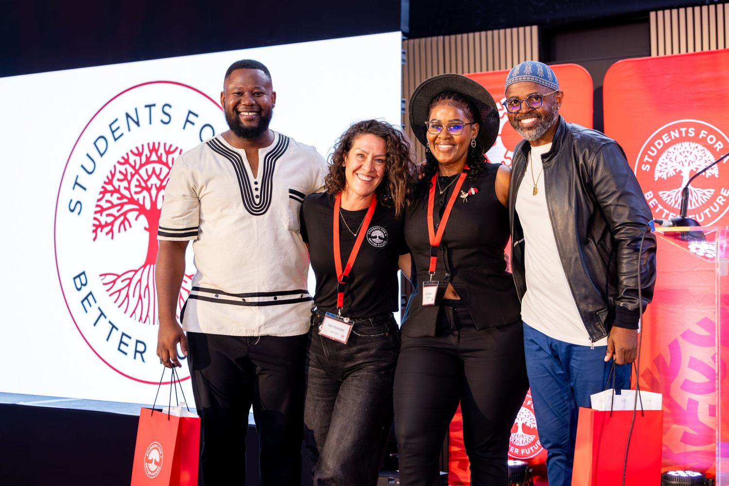 A group of people pose together at an event with red banners and logos visible in the background.