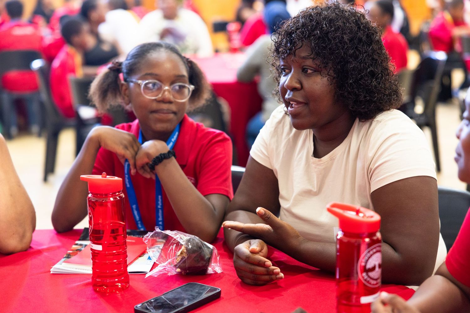 Two people engage in conversation while sitting at a table with red cups and tablecloth at a social gathering.
