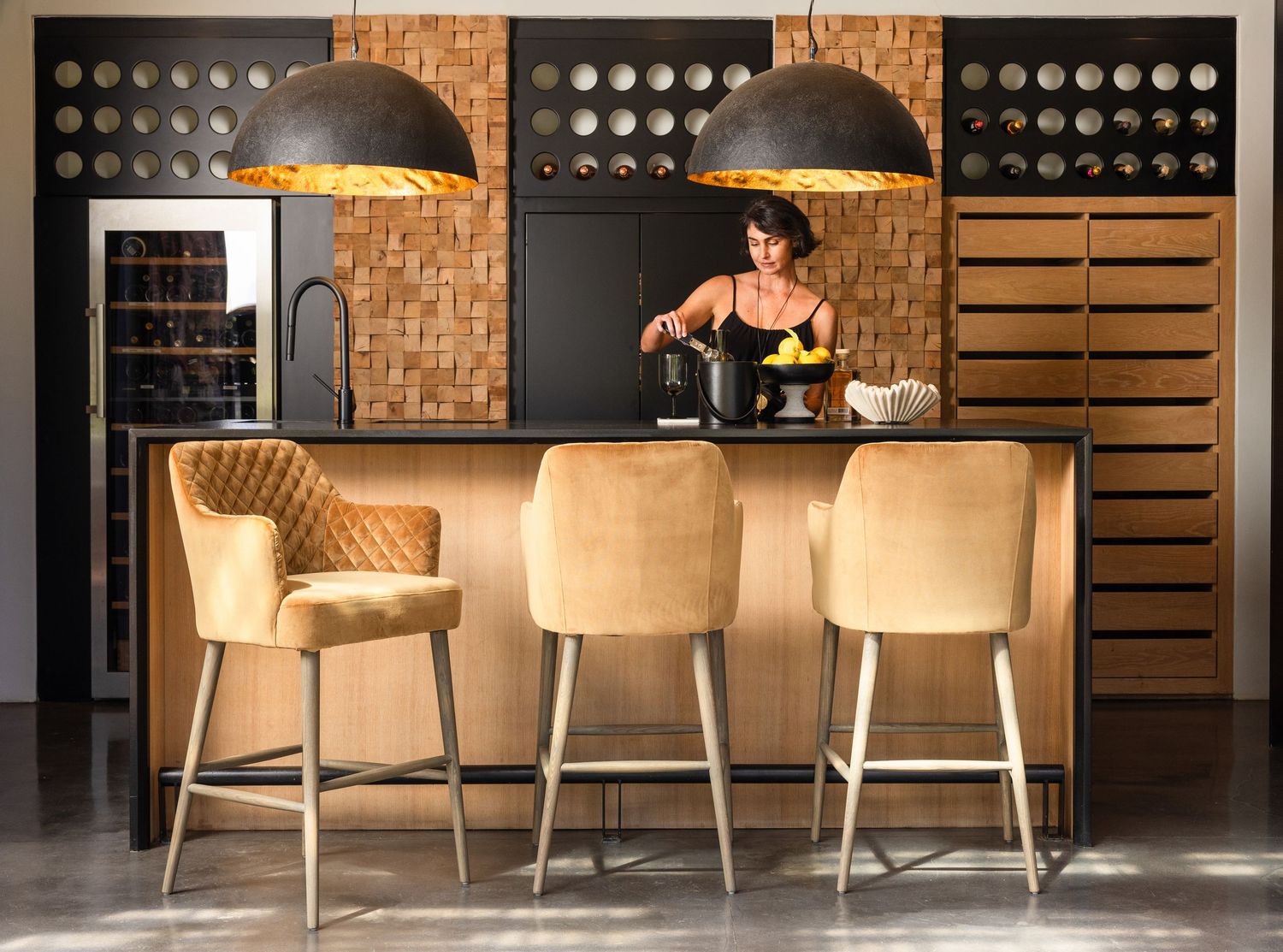 Modern industrial kitchen with wooden bar counter, beige velvet barstools, and pendant lighting in a loft-style setting.