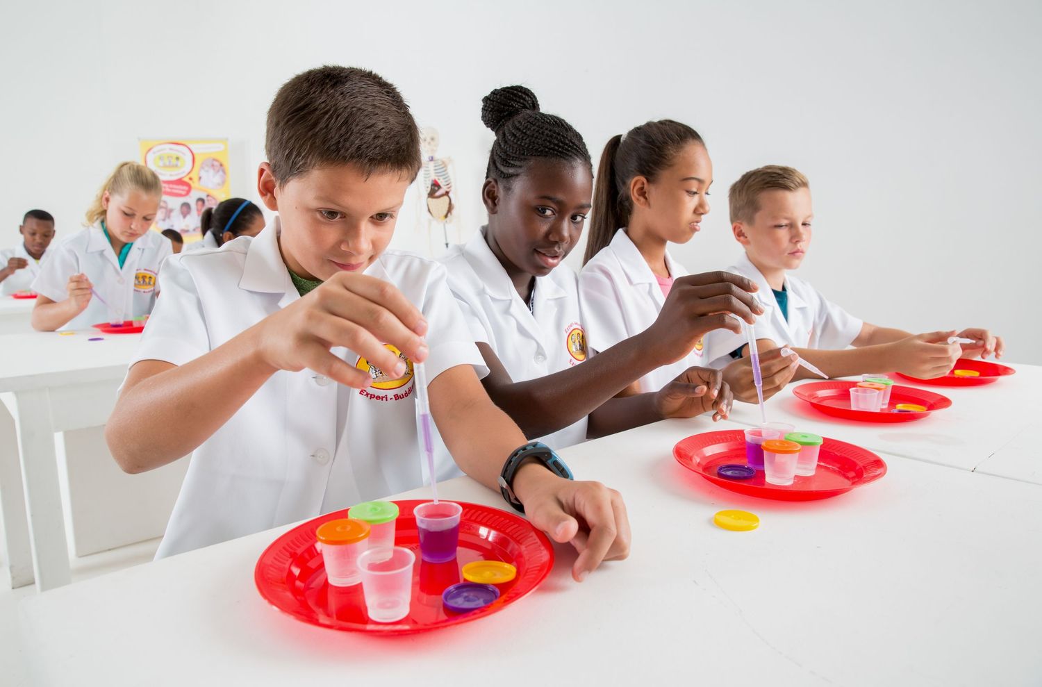 Students in white lab coats work together on science experiments with red trays and test tubes.