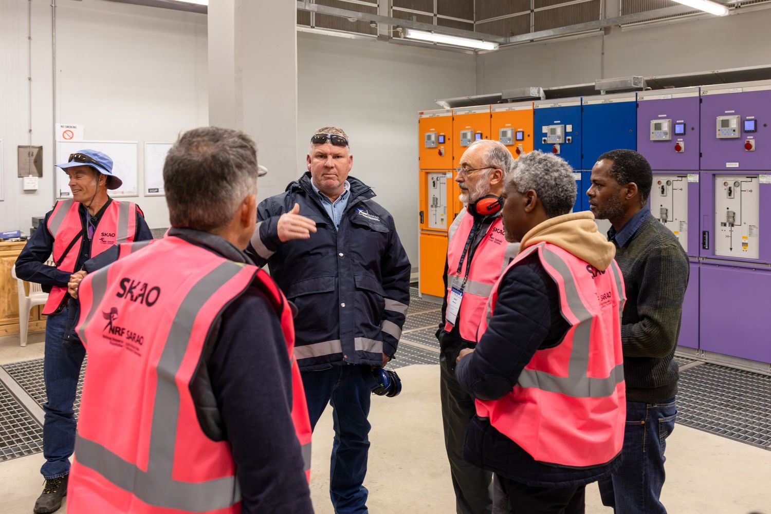 Workers in high-visibility vests having discussion in industrial facility hallway.
