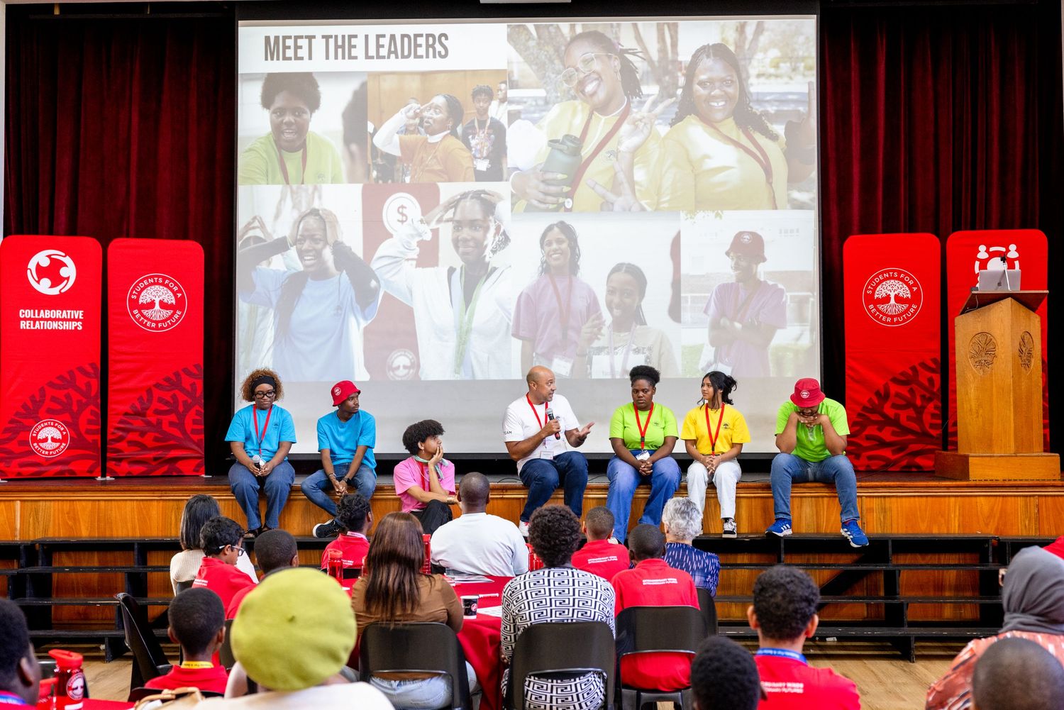 Audience members seated in a theater auditorium with red curtains and projection screens displaying presentations.