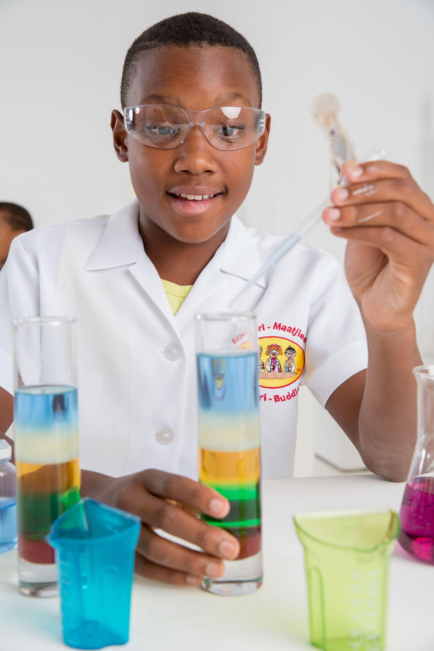 Student wearing safety glasses examines colorful liquid density layers in test tubes during science class.
