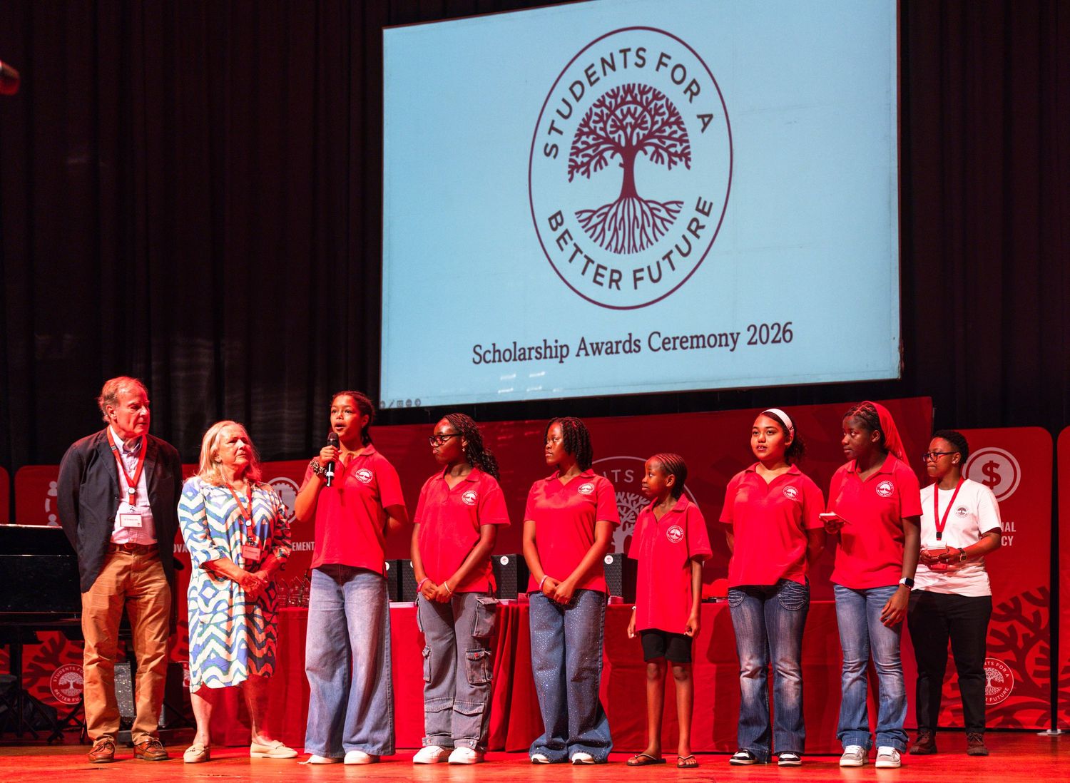 Students in red shirts stand on stage during an awards ceremony with organizational logos displayed on screen.