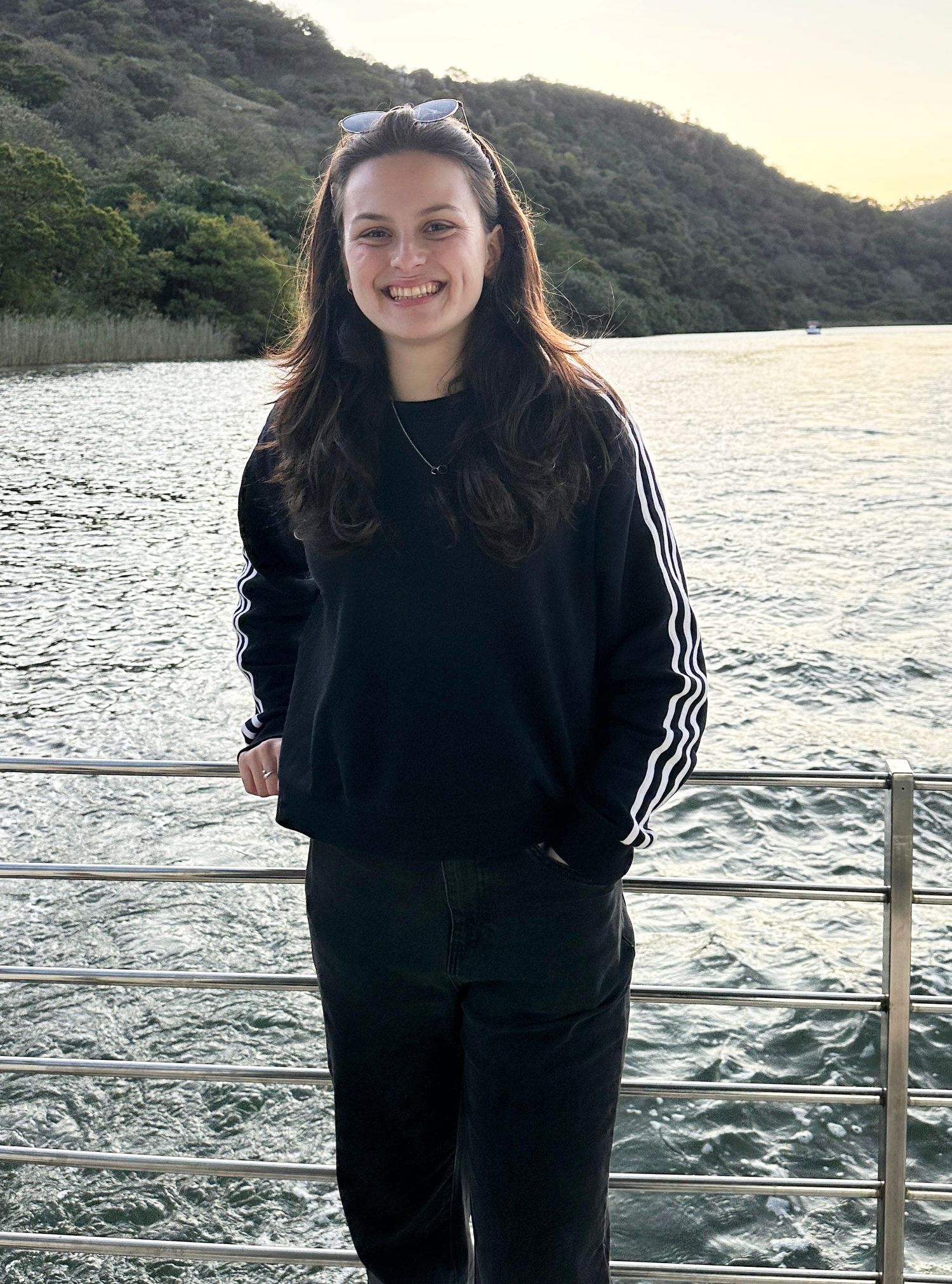 A person in black athletic wear stands on a boat deck against a scenic waterfront and mountain backdrop at sunset.