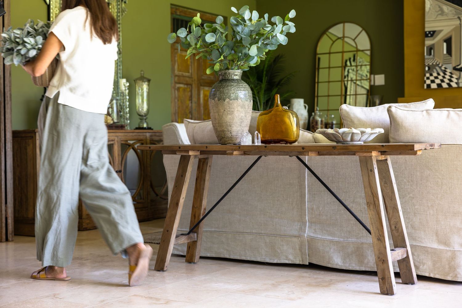 Rustic wood console table with white vase and decorative items against olive green wall in casual living room interior.