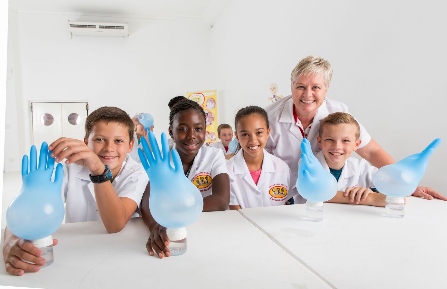 Students and teacher in lab coats show blue gloves during science class.