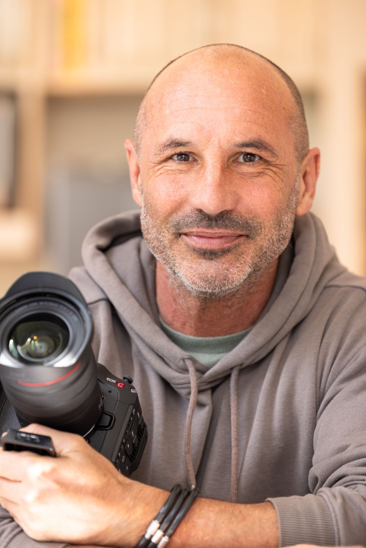 A photographer wearing a gray hoodie holds a professional camera with a telephoto lens while smiling warmly.