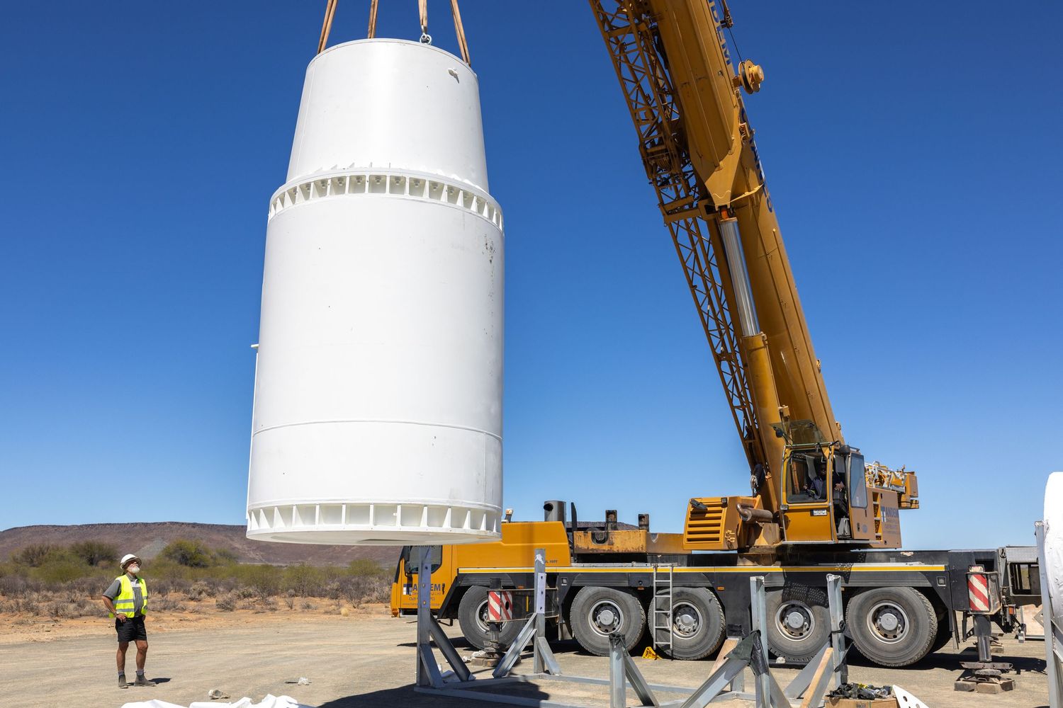 Construction crane lifting large white cylindrical structure at desert work site.