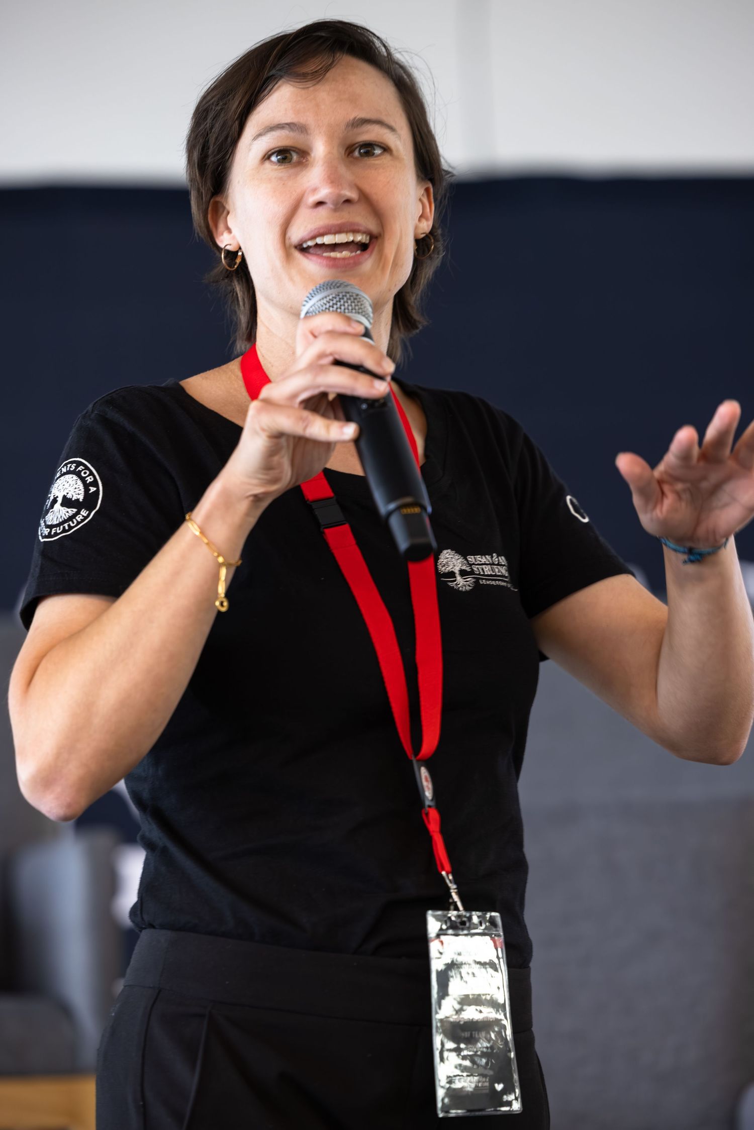 Speaker with microphone presents on stage wearing black uniform and red lanyard at indoor event.
