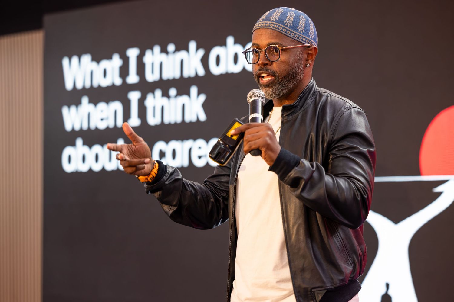 A speaker wearing a black jacket and glasses gestures on stage during a presentation with text displayed behind them.