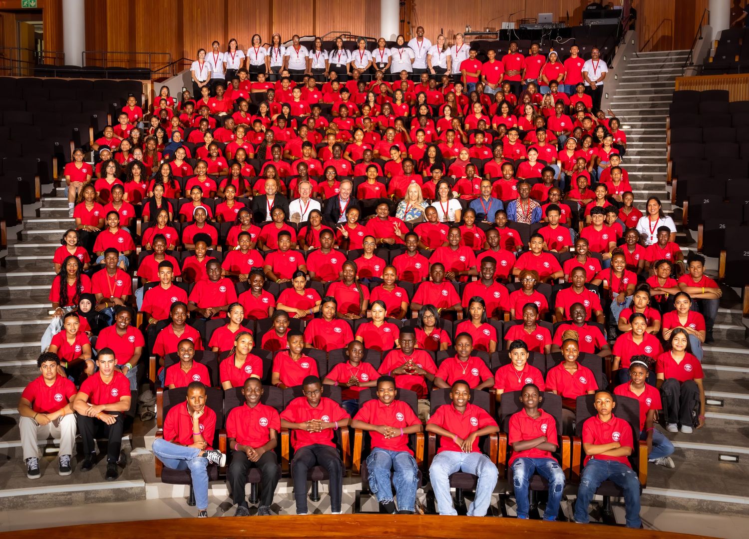 Large group photo of graduates wearing red shirts seated in rows in an auditorium setting.
