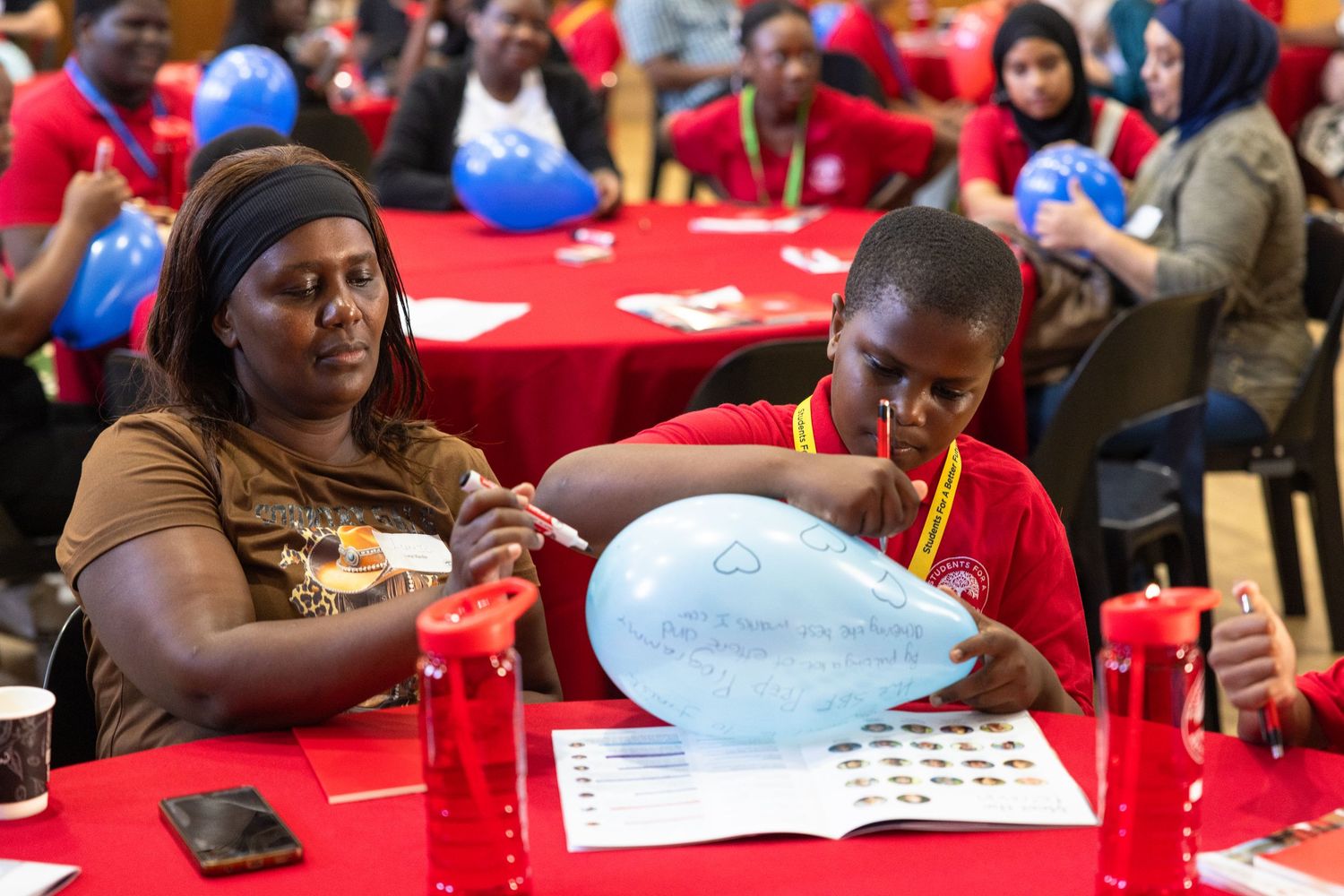 Students sit at red tables working on papers and activities during an educational workshop session.