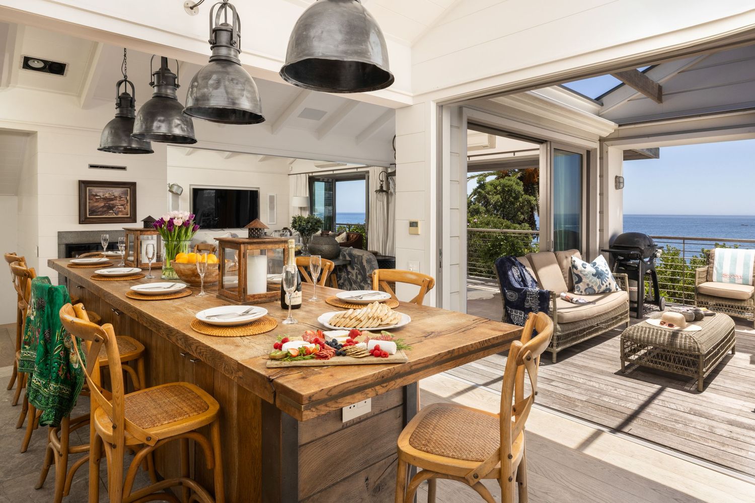 Modern coastal kitchen and living room with ocean views features wooden dining table and industrial pendant lights.