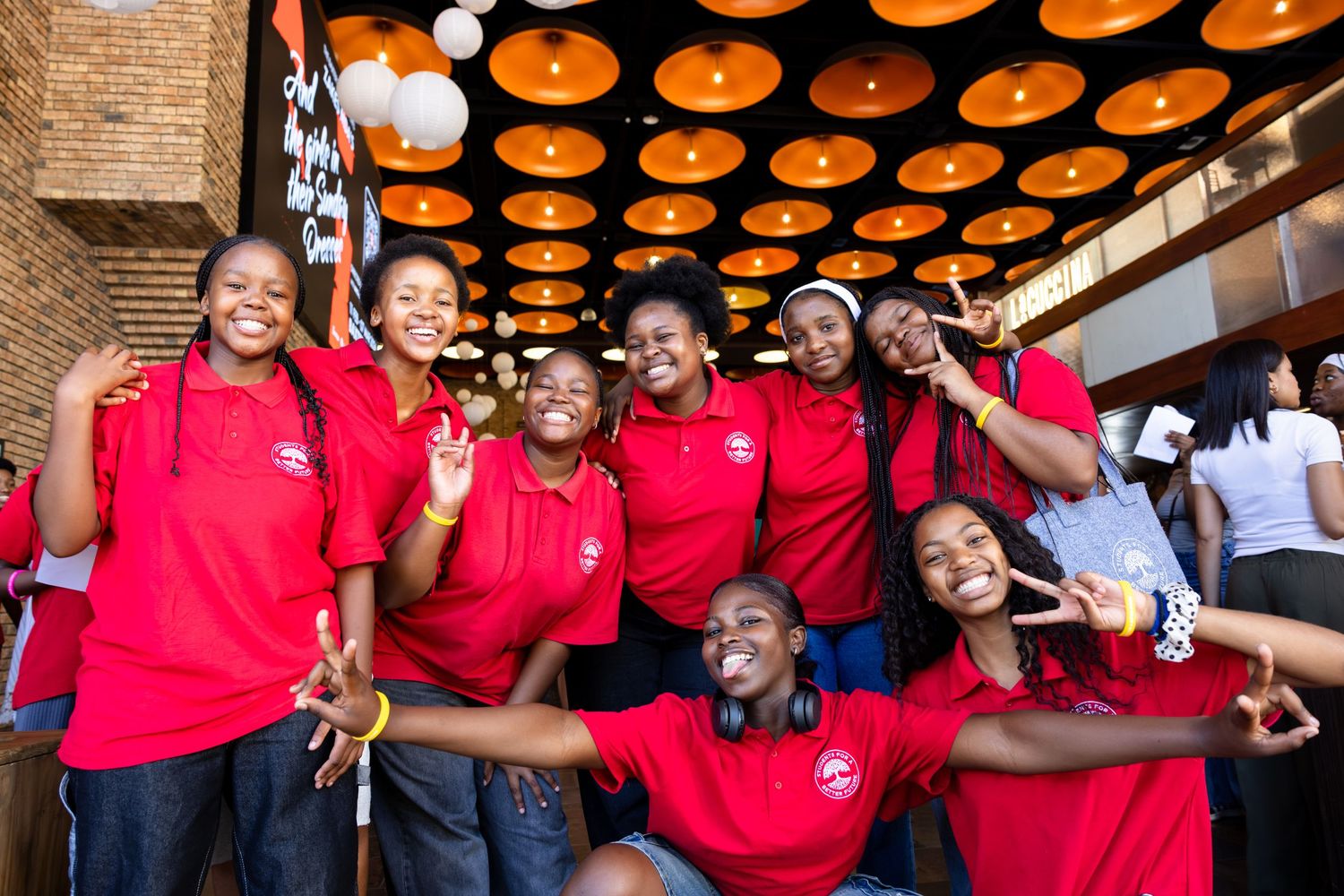A group of restaurant staff in matching red polo shirts pose together under decorative orange ceiling lights.