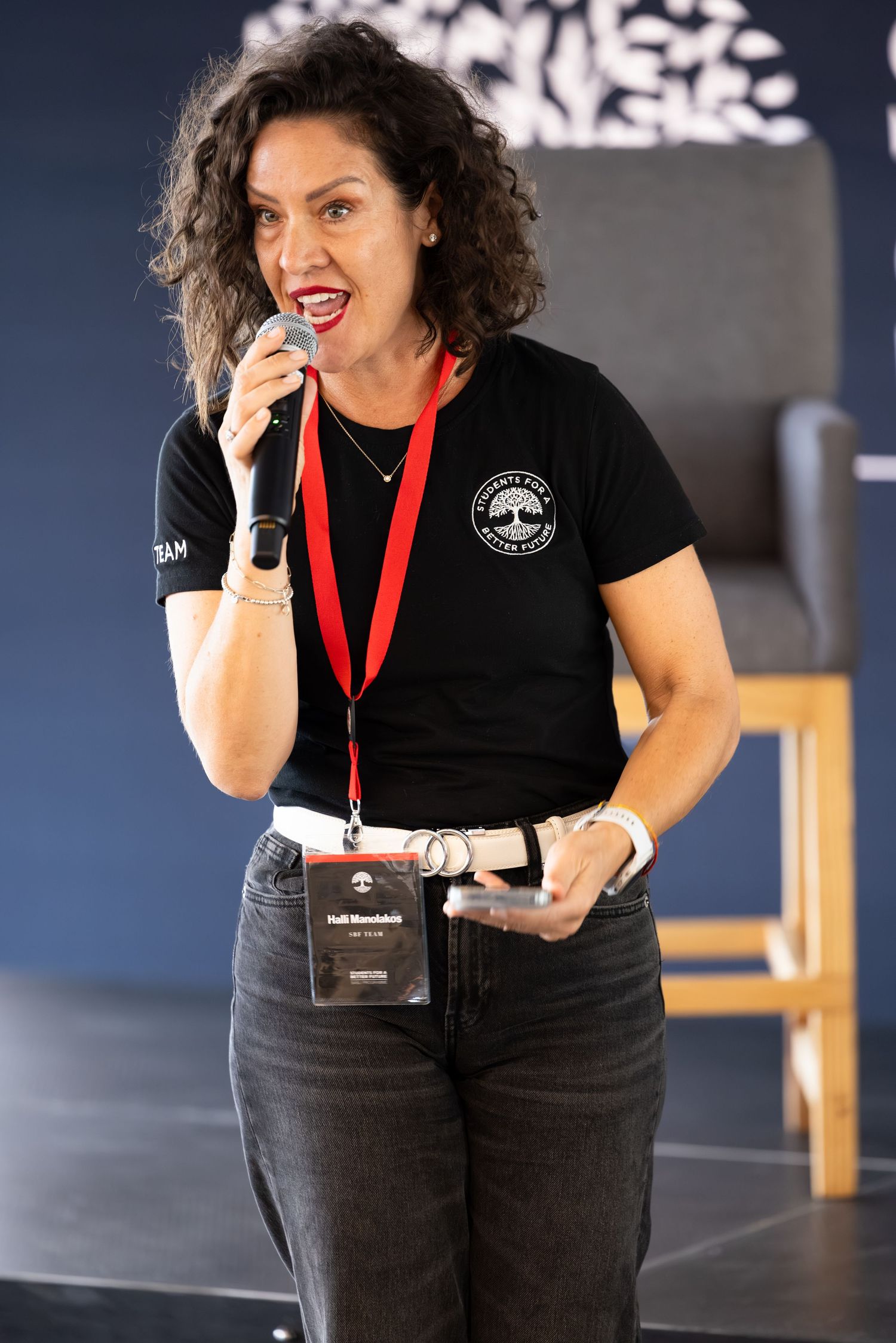 Event presenter in black t-shirt speaks into microphone against dark backdrop with event branding.