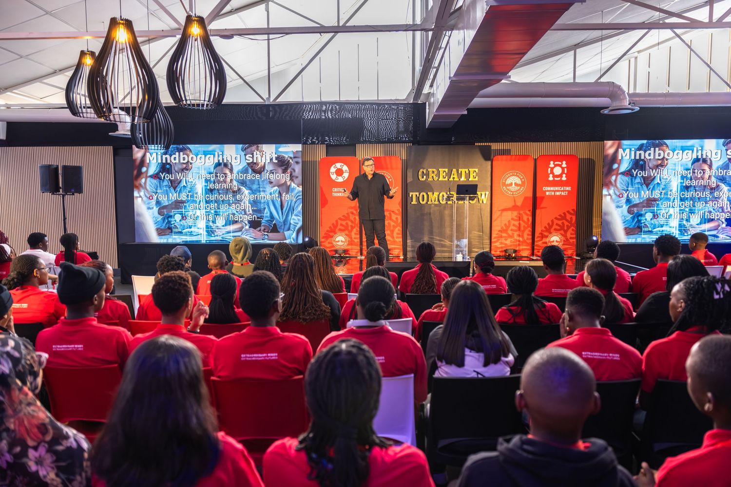 A large audience in red shirts watches a presentation on big screens at a corporate event or conference.