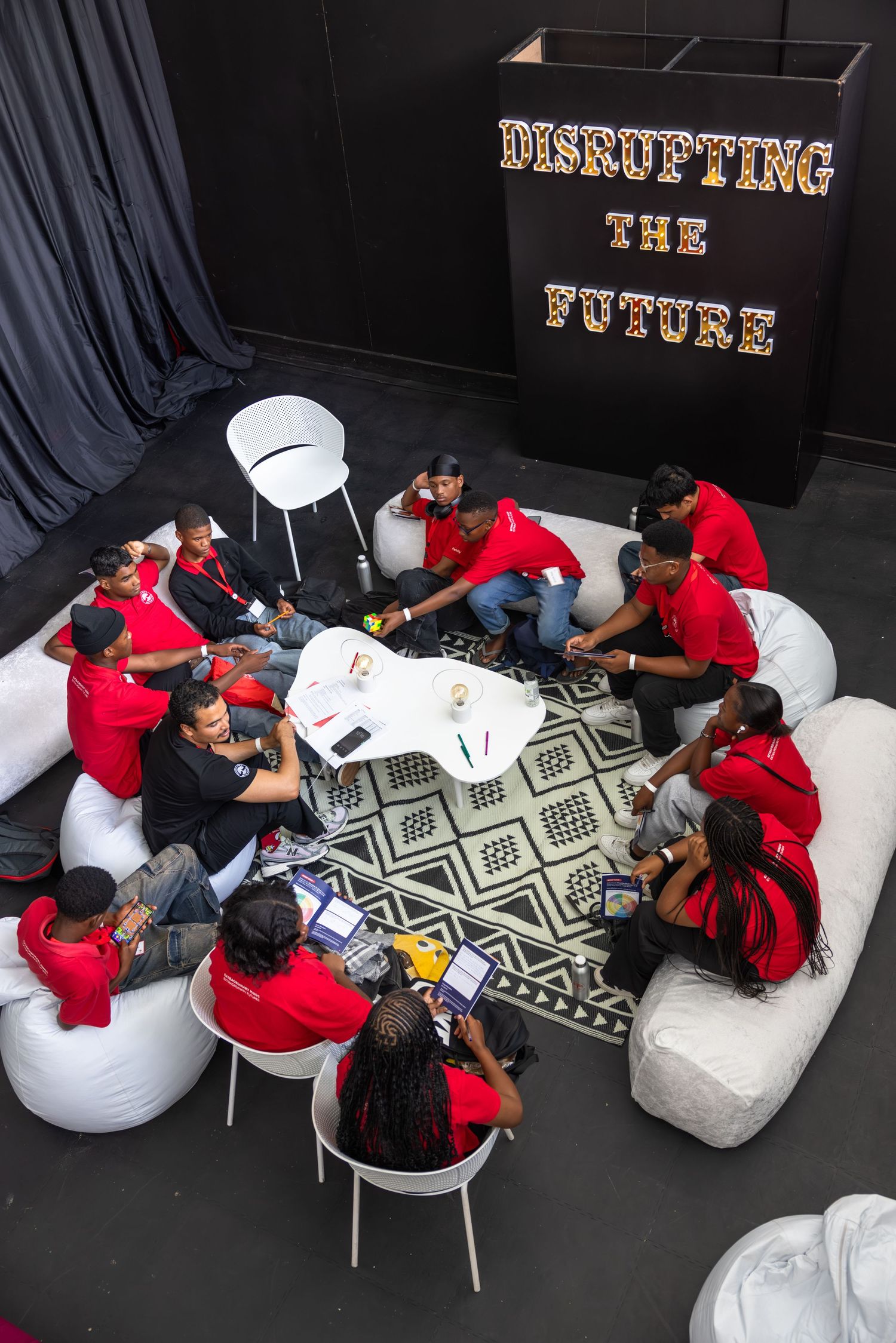 Students in red shirts gather around a table for a collaborative session under a sign reading 'Disrupting the Future'.