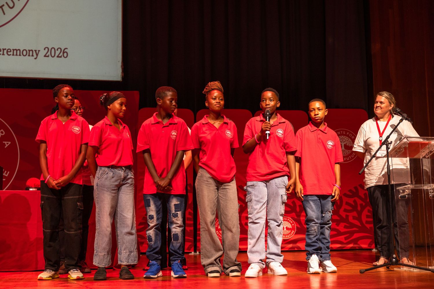 Students in red shirts and gray pants perform together on a brightly lit stage during a school event.