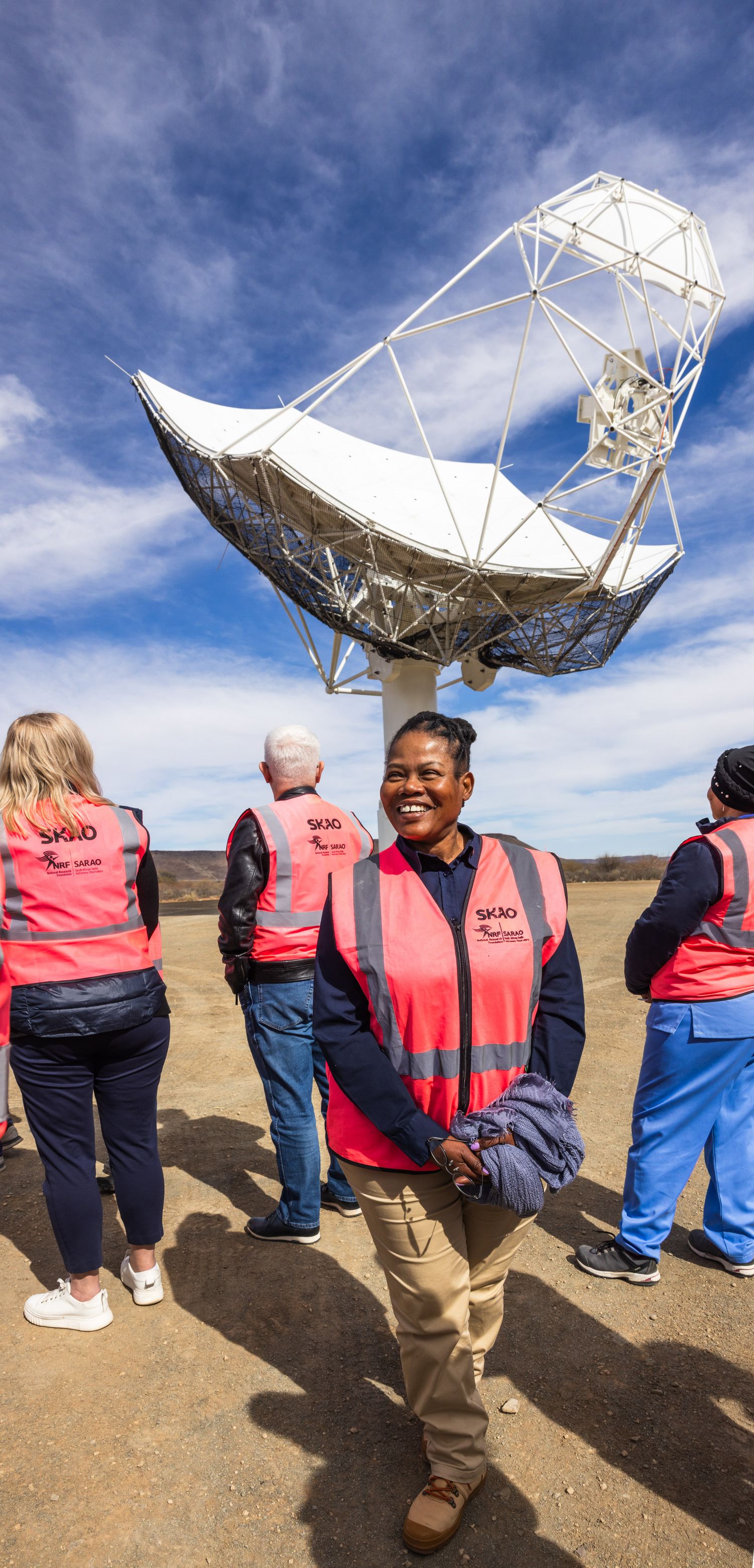 Tour group wearing safety vests stands beneath a large white radio telescope dish against blue sky.