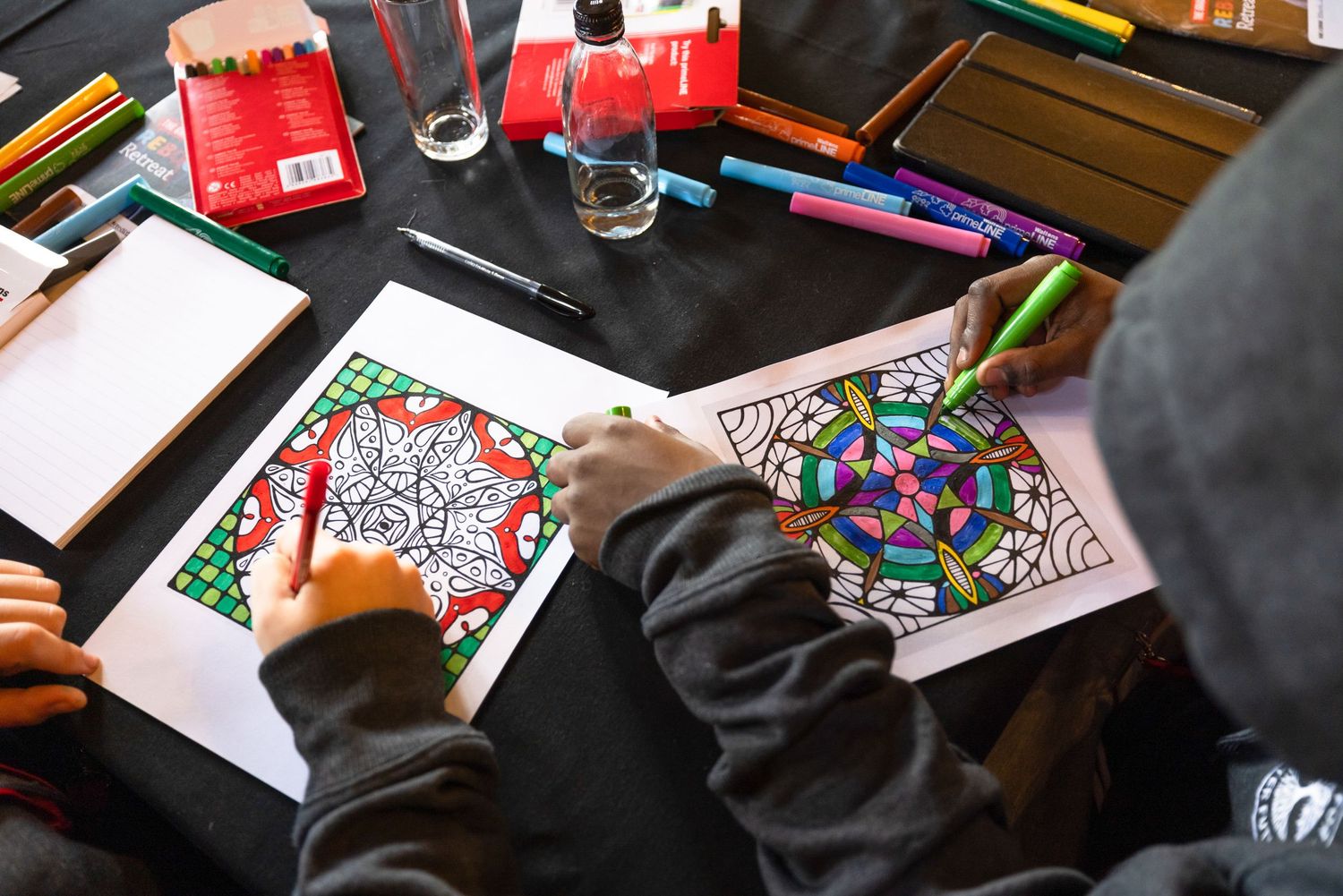 Hands working on colorful mandala artwork with markers on black surface.