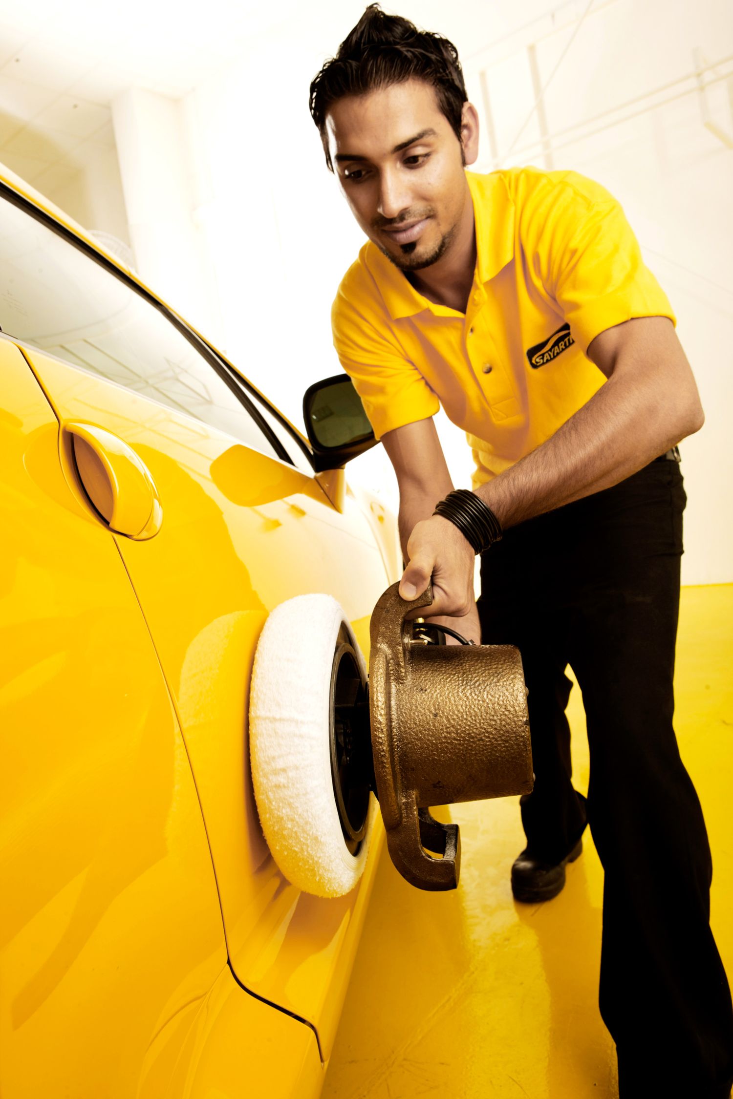 Auto service technician in yellow uniform polishing yellow car exterior at service center.