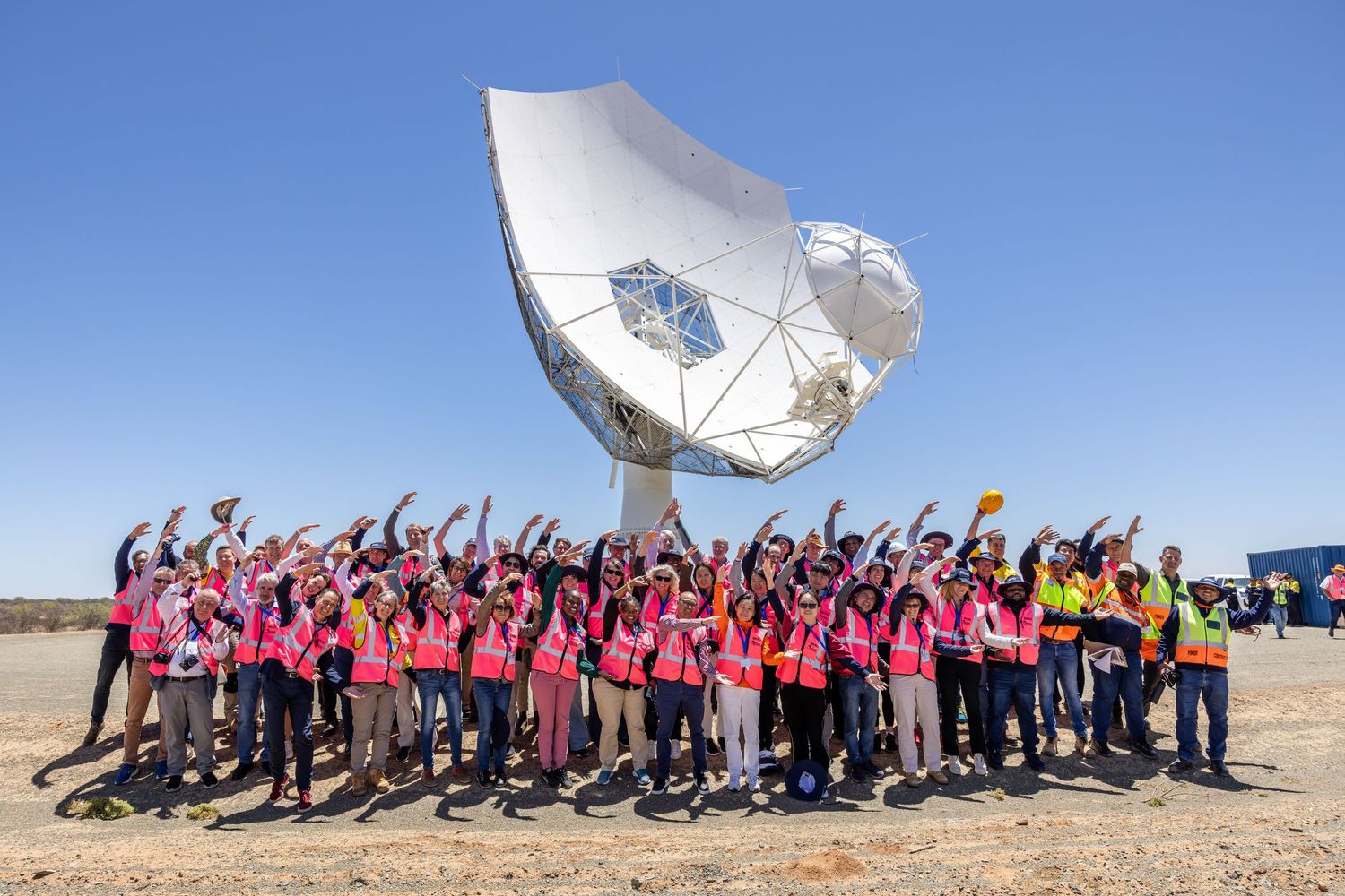 Large group poses in front of satellite dish wearing matching pink shirts.
