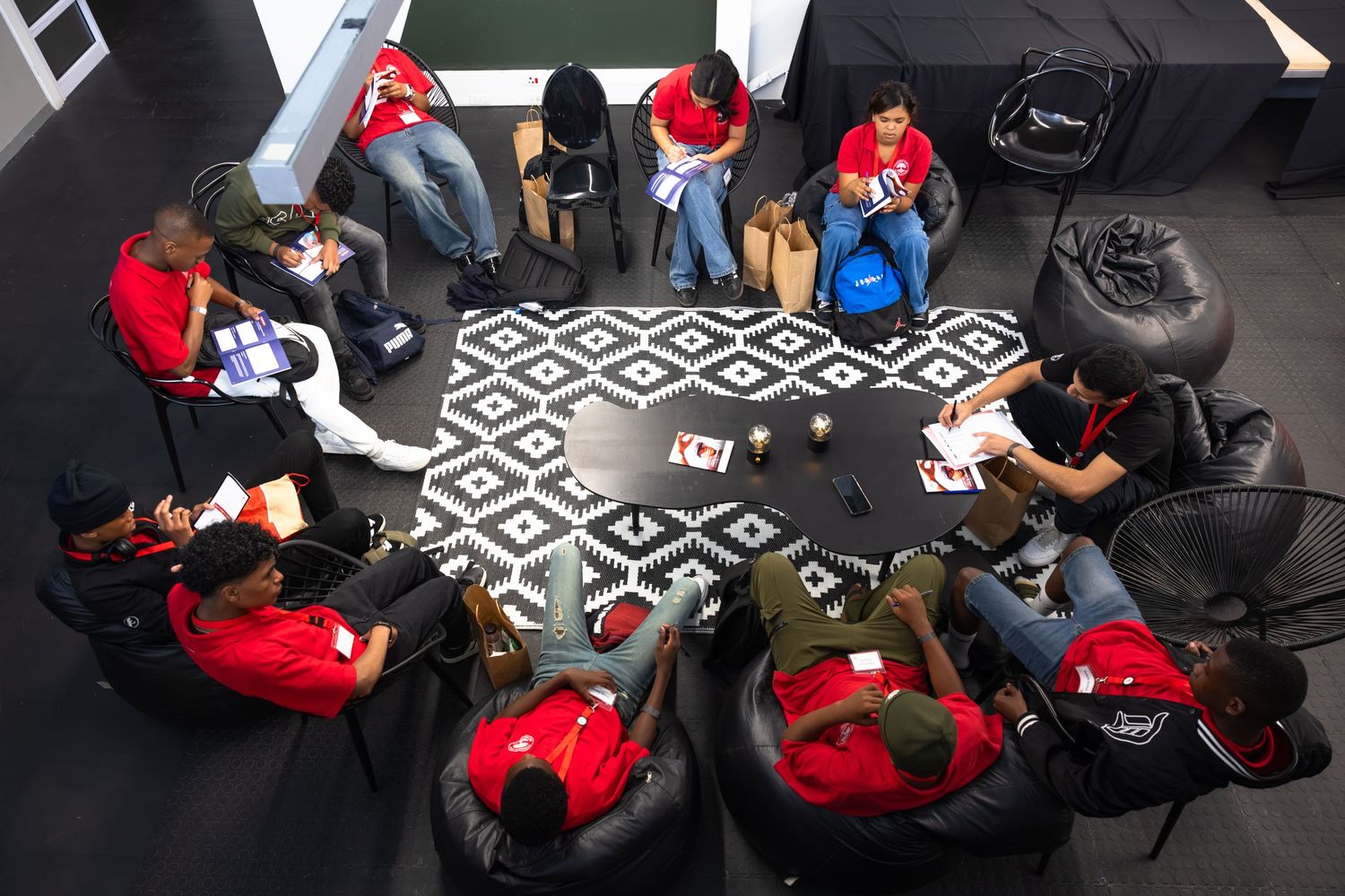 Students in red uniforms sit at tables with black and white geometric pattern carpeting during a group activity.