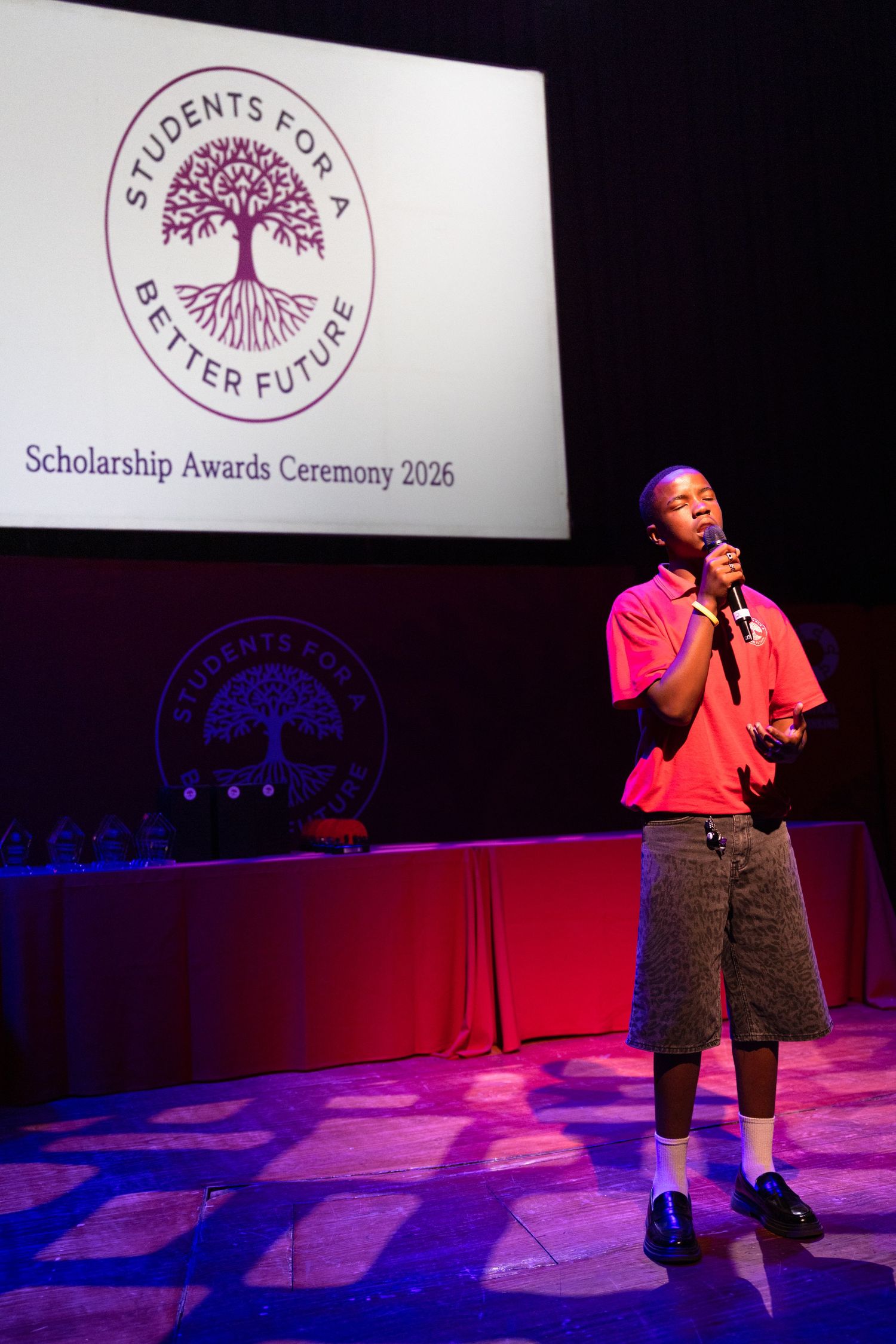 A performer in a red shirt speaking on stage during a scholarship awards ceremony.