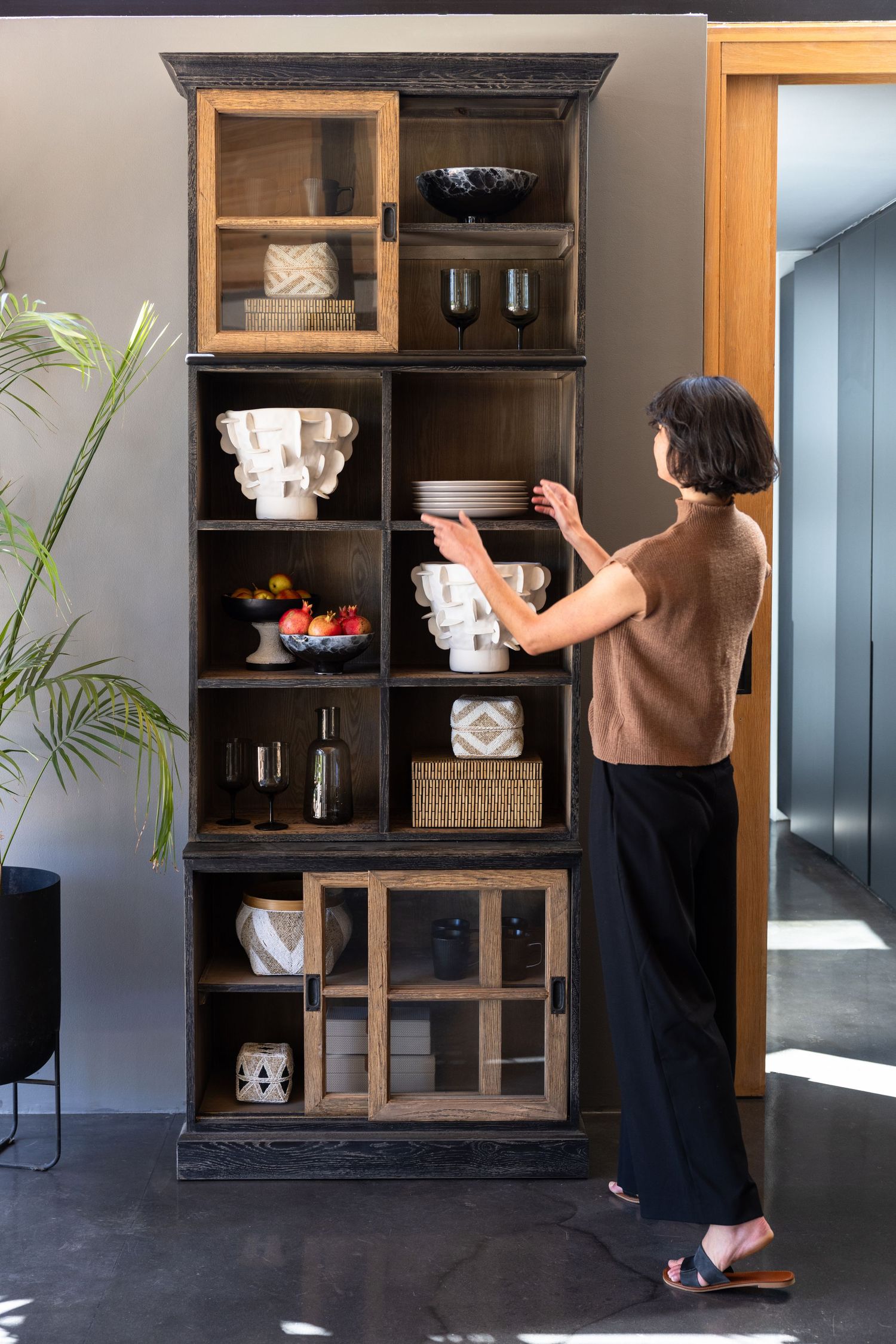 A person arranges decorative items on a tall black wooden shelving unit with multiple compartments in a modern interior space.