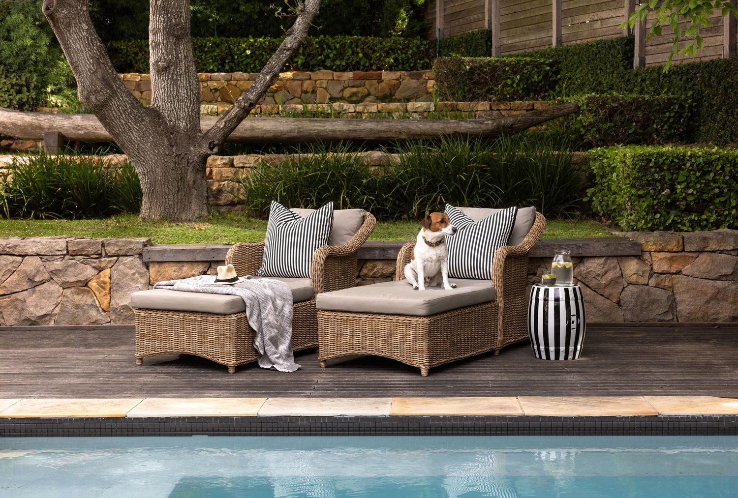 Outdoor wicker lounge chairs line a swimming pool deck with stone walls and landscaping in the background.