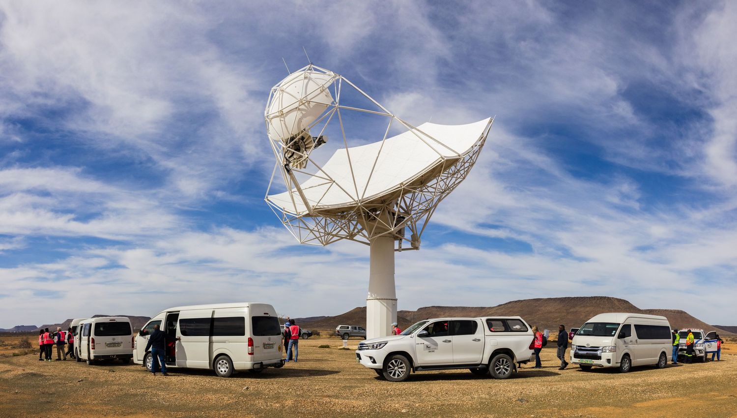 White satellite dish installation with vehicles parked nearby against desert landscape.