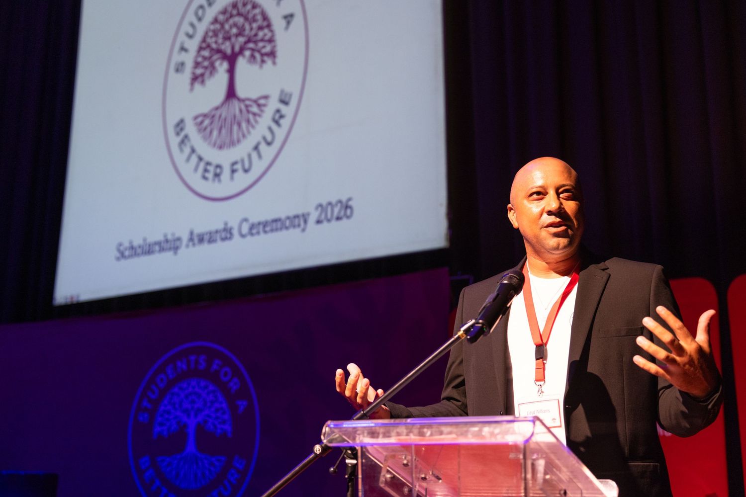 A speaker presents at an awards ceremony with a large projection screen displaying a logo in the background.