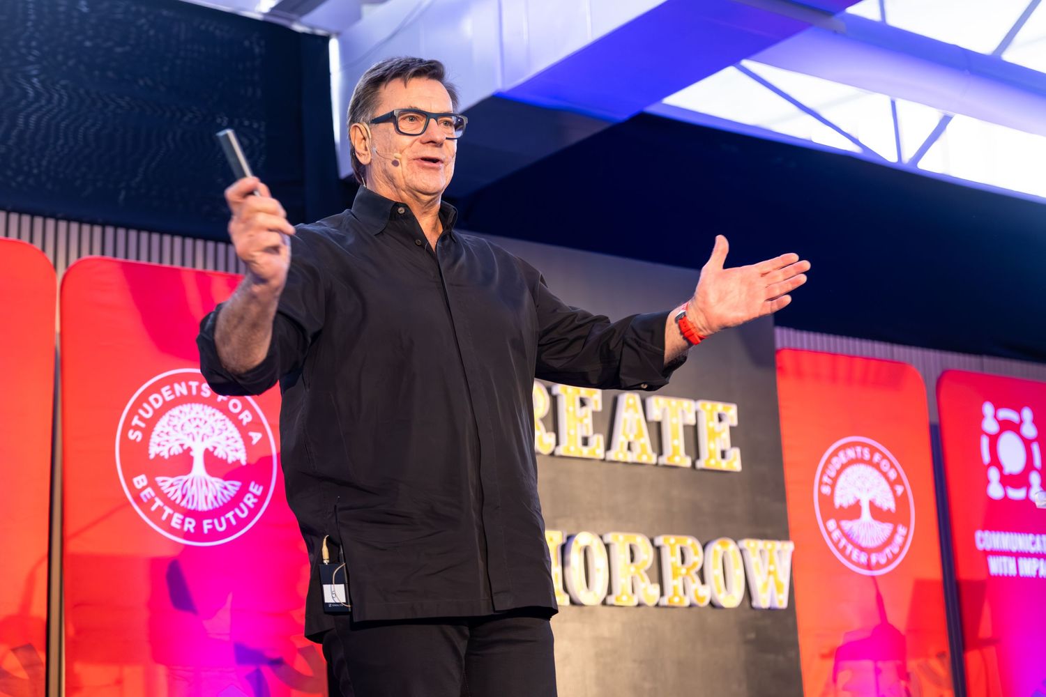 Speaker presents on stage with large digital display screens and red branding elements.
