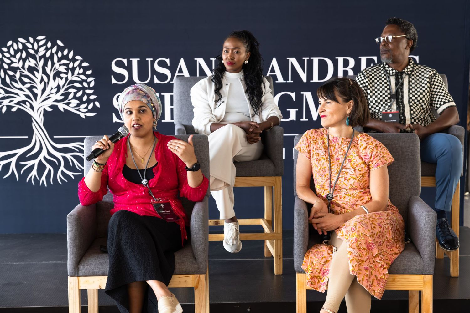 People seated in front of a dark wall with text and tree designs during an event.