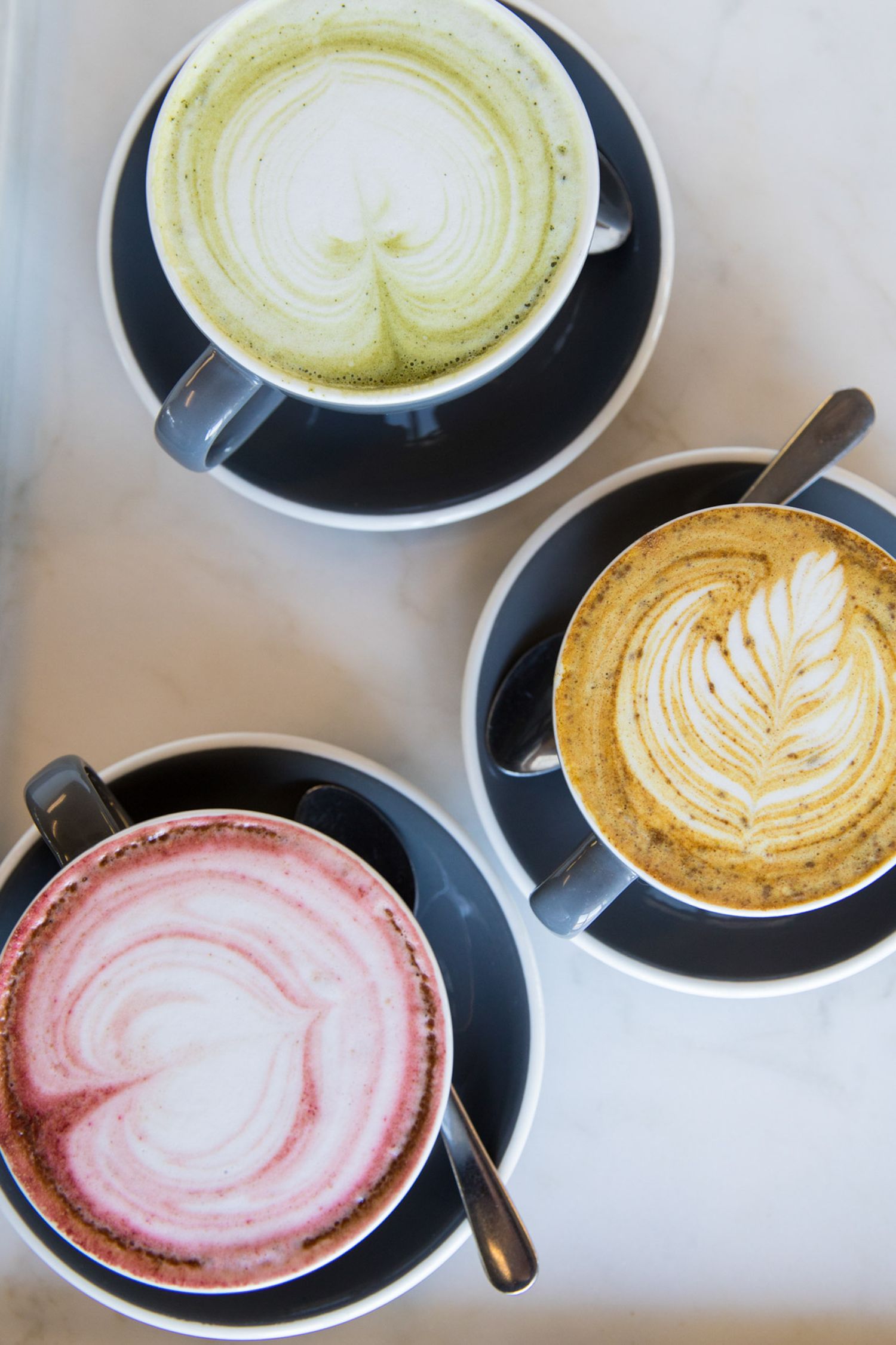 Three artistic latte drinks featuring pink, green, and golden foam art designs.