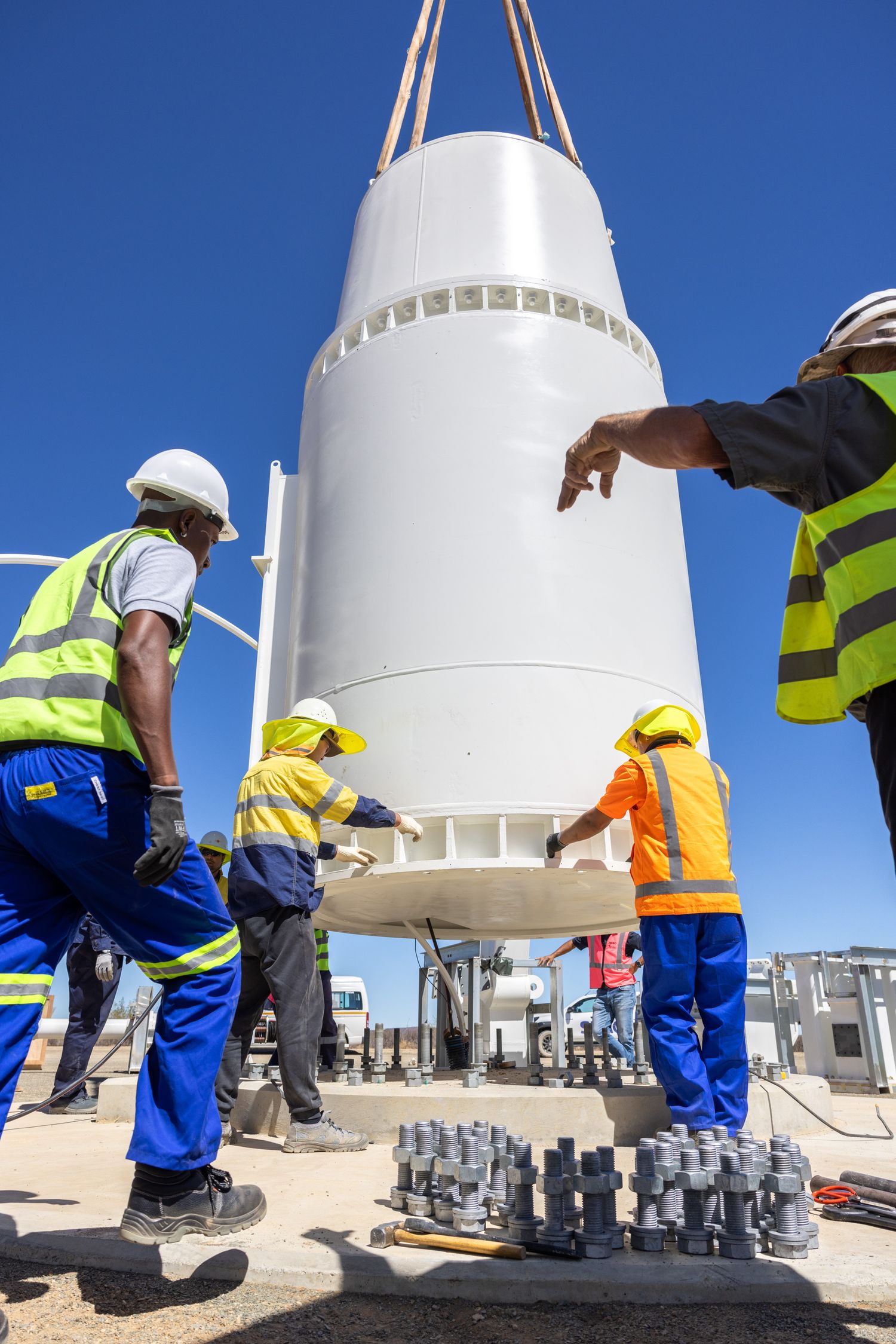 Workers in safety gear assemble components of a large white industrial structure against a bright blue sky.