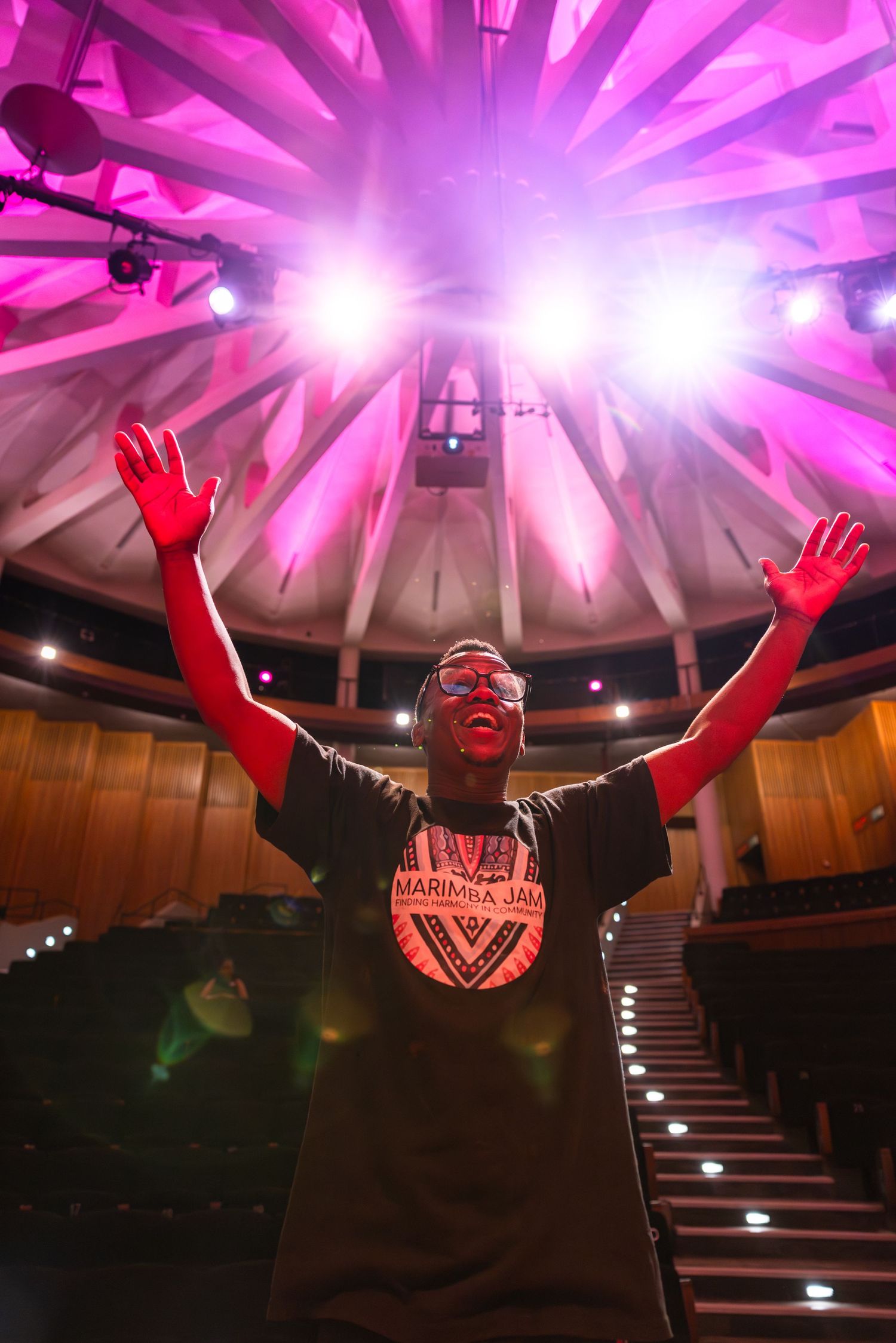 A performer stands with arms raised in celebration under dramatic pink stage lighting in an auditorium.