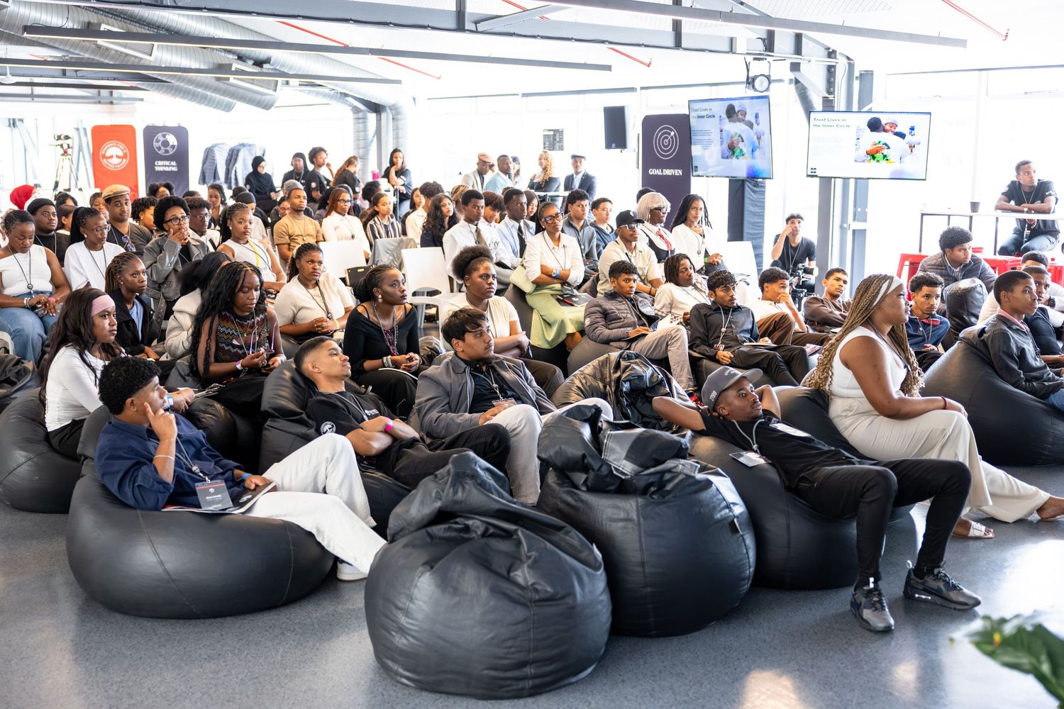 Large audience seated in bean bag chairs attentively watches presentation in modern conference space.