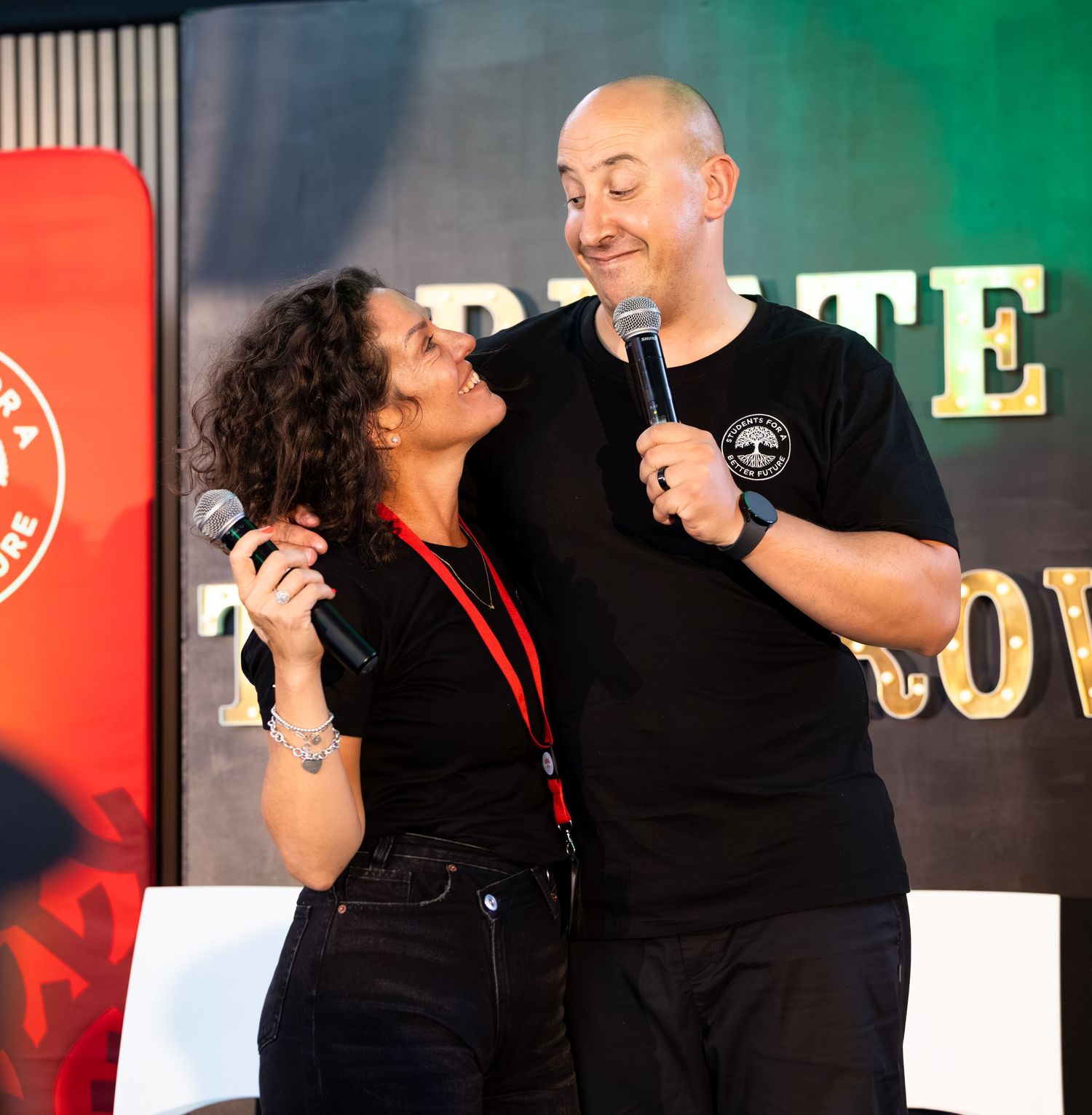 Two speakers share microphones while presenting on stage with dark backdrop.