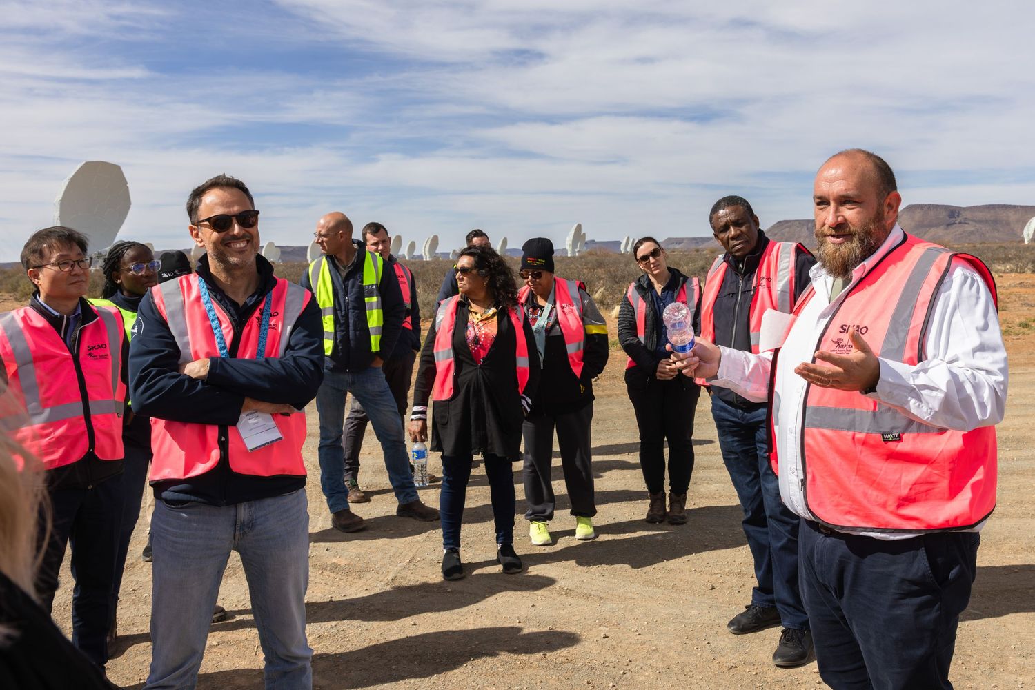 Group of visitors in safety vests touring a radio telescope facility in a desert setting.