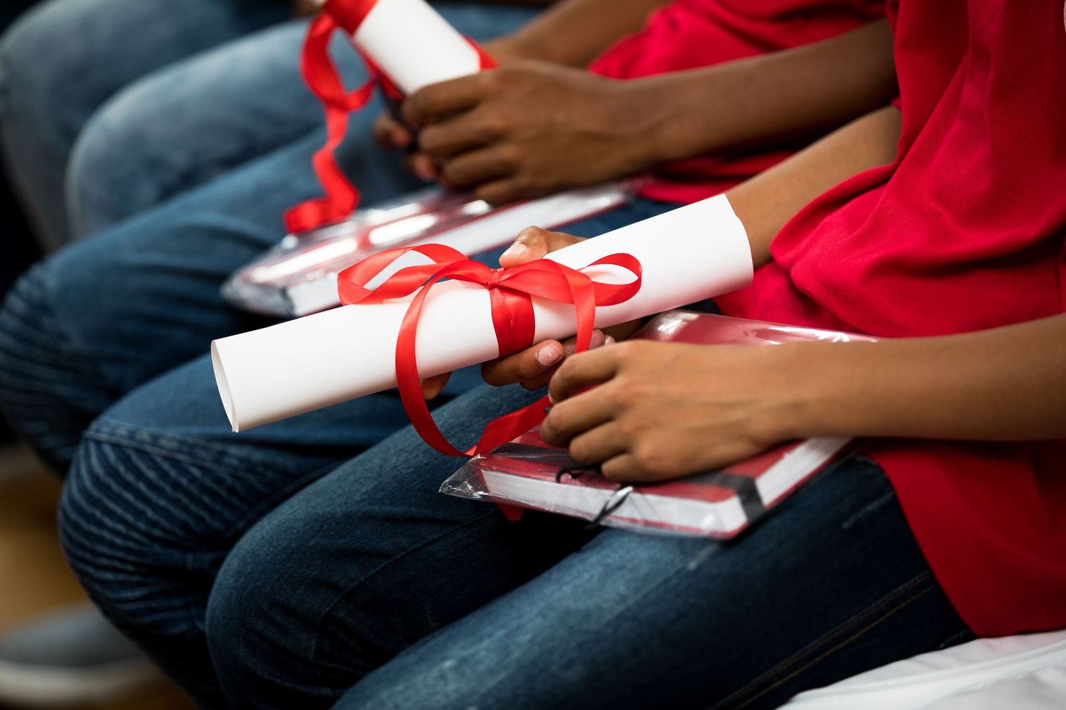 Hands holding rolled diplomas with red ribbons while sitting in jeans.