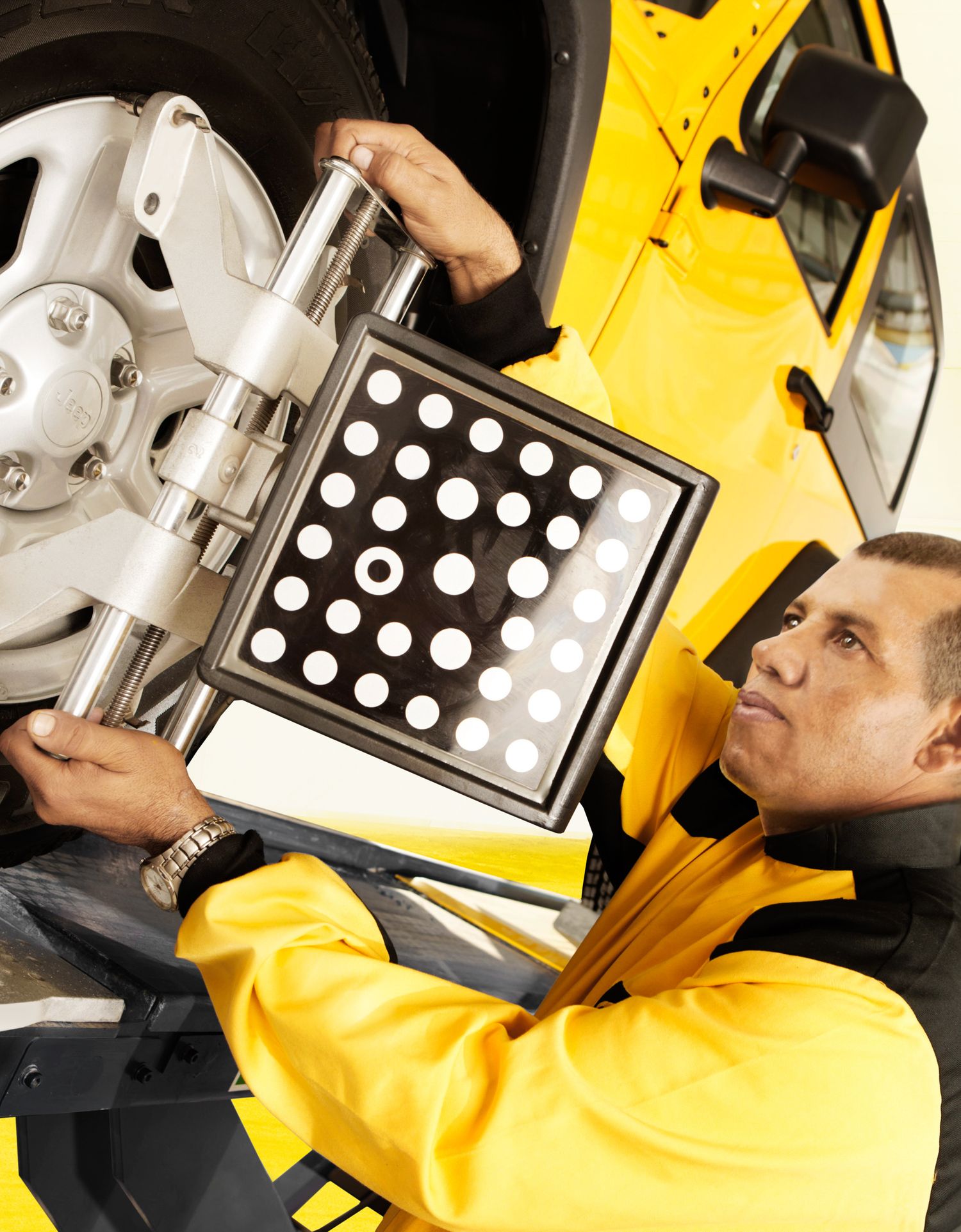 Auto technician performs wheel alignment and checks suspension using computerized diagnostic equipment in yellow uniform.