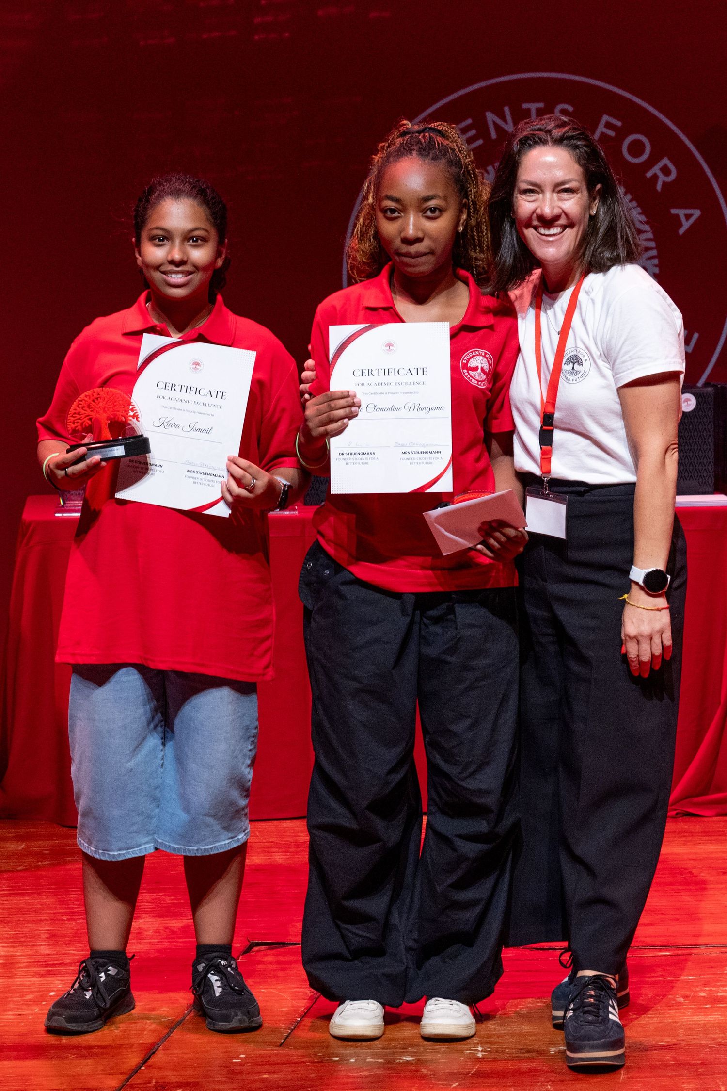 Students in red shirts and black pants proudly display their certificates during a graduation ceremony.